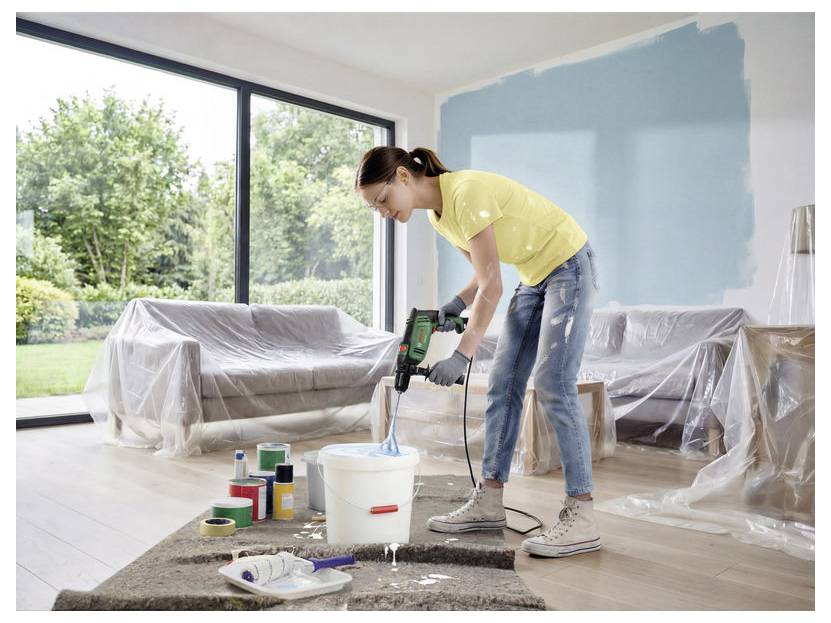 Vrouw die een muur schildert met een boor-aangedreven mixer in een heldere woonkamer die wordt gerenoveerd, met meubels afgedekt met plastic en verfbenodigdheden op de vloer.