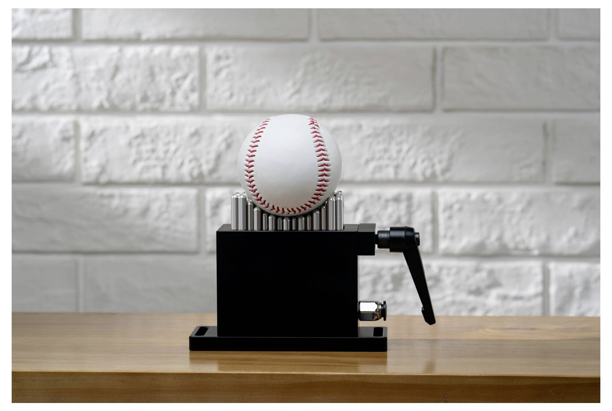 Baseballnaaimachine met een honkbal gemonteerd op een zwarte handmatige pers op een houten tafel tegen een witte bakstenen muur.