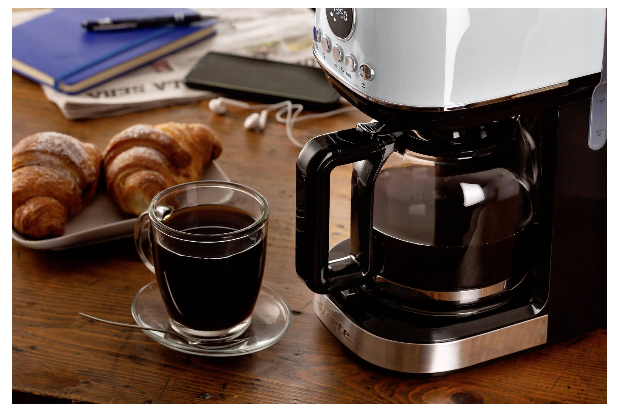 Koffiezetapparaat dat koffie brouwt op een houten tafel naast een glazen kopje en croissants voor het ontbijt.