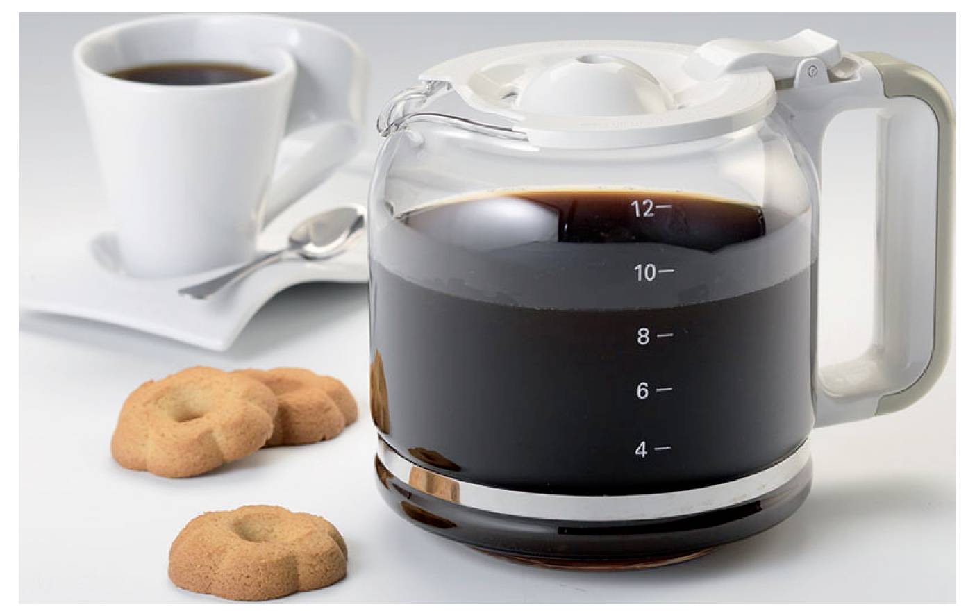 Glazen koffiepot gevuld met gezette koffie naast een kopje en schotel, met koekjes op de voorgrond.