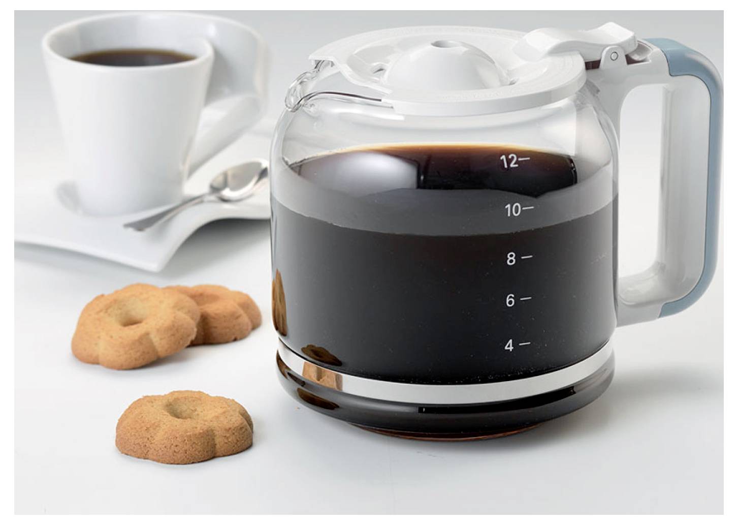 Een glazen koffiepot gevuld met zwarte koffie staat naast een kopje, lepel en koekjes op een wit oppervlak.