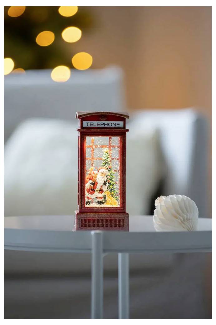 Kleine rode telefooncel-ornament met een kerstman-figuur erin, gloeiend op een tafel in een zacht verlichte kamer met onscherpe lichten erachter.