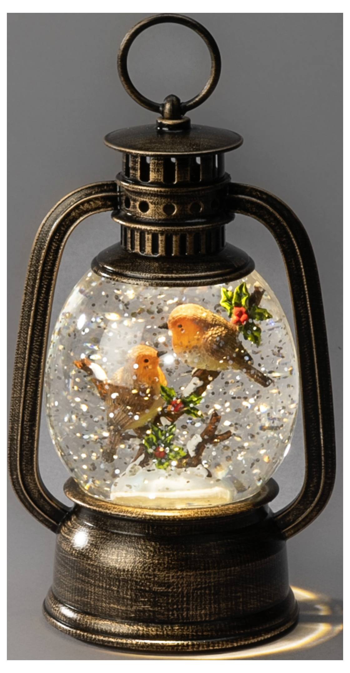 Decoratieve lantaarnvormige sneeuwbol met twee oranje vogels, hulst en glitterend sneeuw in een glazen kom op een donkere metalen voet.