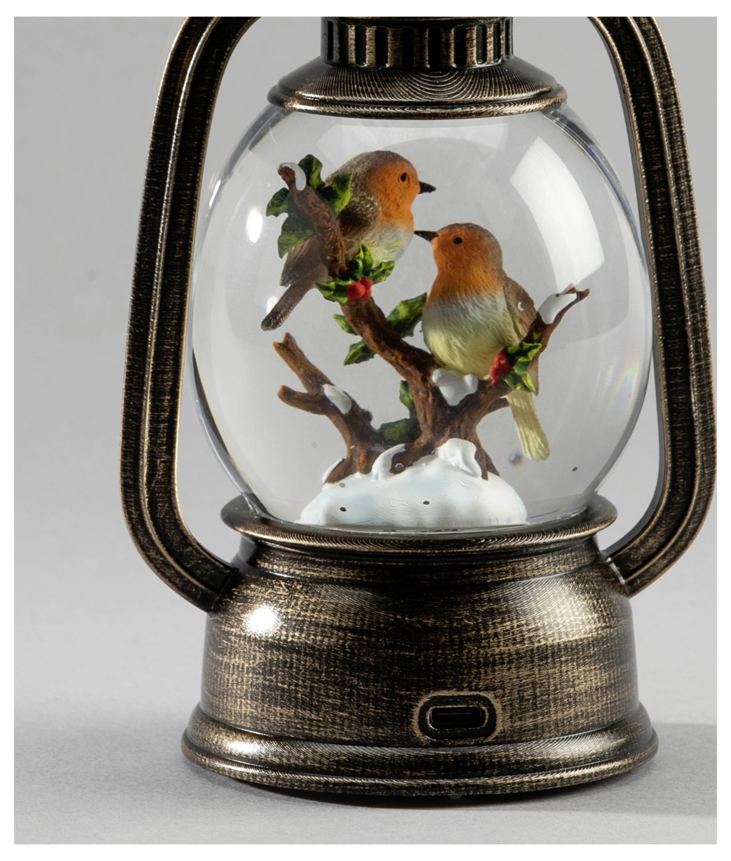 Een decoratieve lantaarn met twee kleine oranje vogeltjes op takken die op een glazen bol neerkijken, van onderaf verlicht.
