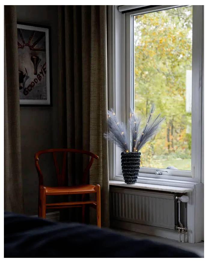 Een houten stoel staat naast een raam met een vaas met verlichte decoratieve takken, met uitzicht op herfsttomen buiten.