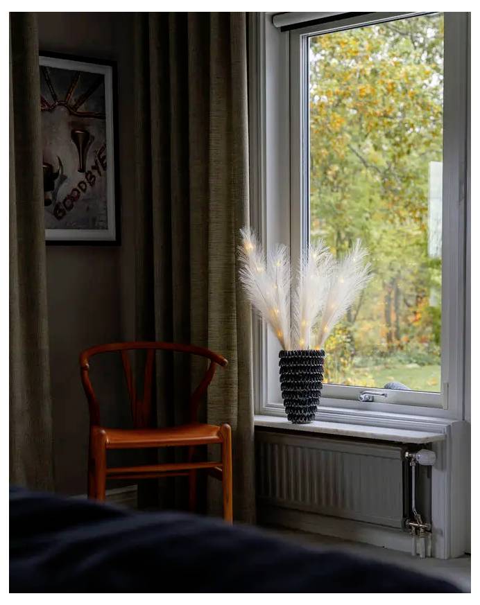 Een houten stoel staat naast een raam met een vaas met witte decoratieve takken, uitkijkend op herfstbomen buiten.