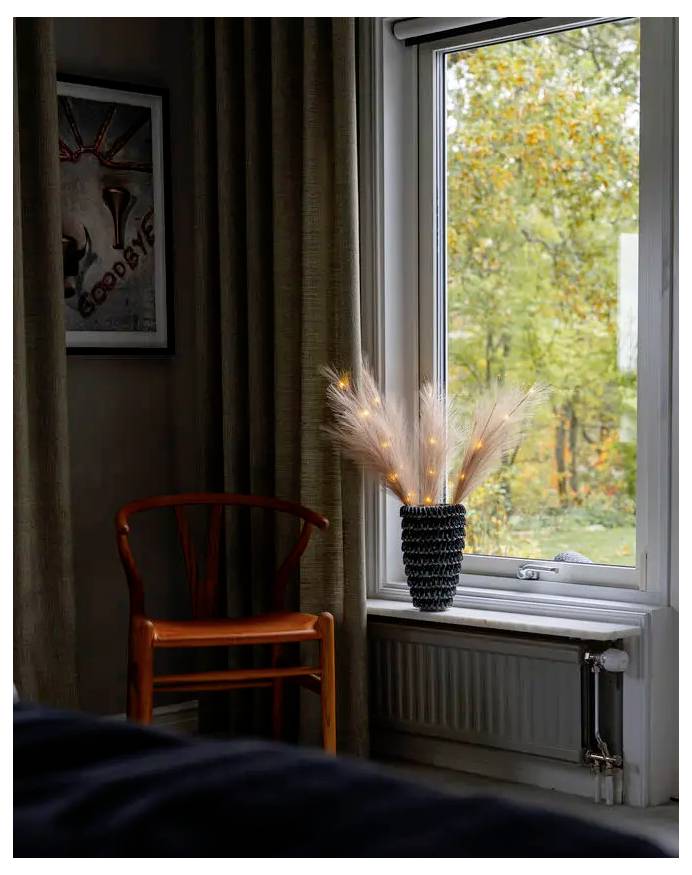 Donkere kamer met een oranje houten stoel bij een raam, een vaas met verlichte decoratieve takken op de vensterbank, en herfsttaken buiten.