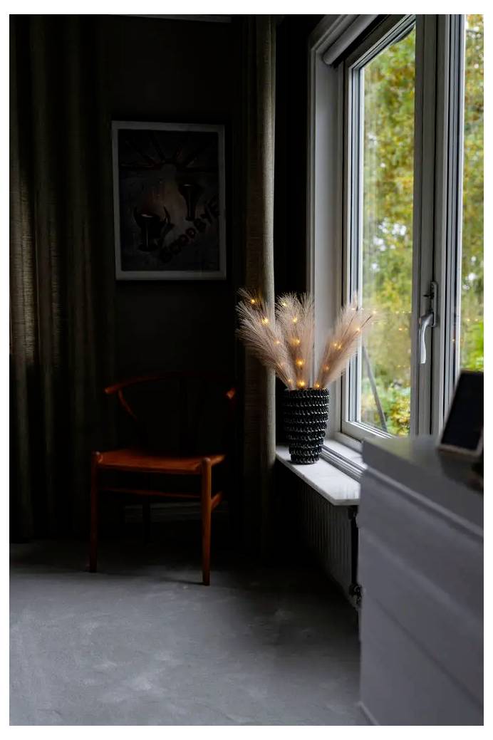 Donker vertrek met een houten stoel tegen de muur en een vaas met verlichte decoratieve takken op de vensterbank naast een openstaand raam.