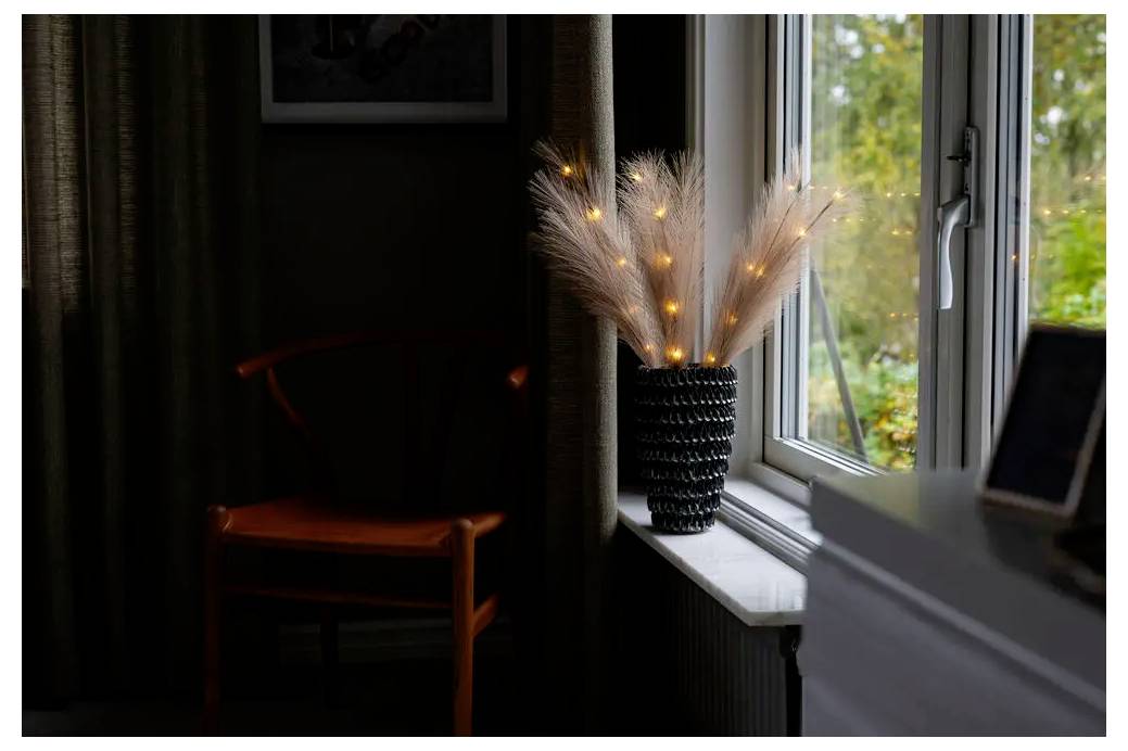 Een verlichte vaas met decoratieve gedroogde stengels staat op een vensterbank naast een openstaand raam, met een donkere houten stoel in het schemerdlige vertrek.