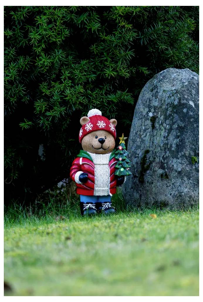 Kleine teddybeerfigurine in een rode wintermuts en jas die een piepklein kerstboompje vasthoudt naast een grote rots in een groen buitenlandschap.
