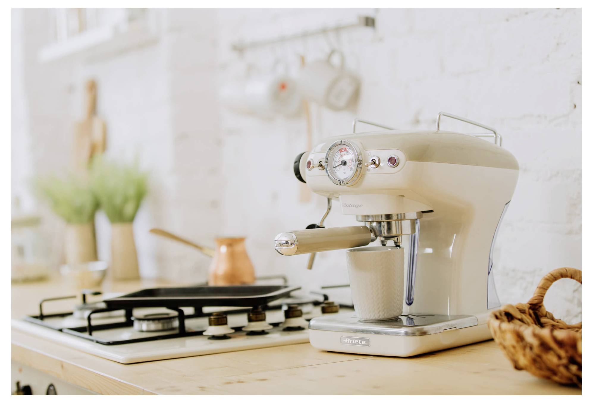 Crèmekleurig espressomachine die een kopje zet op een keukentafel naast een gasfornuis en een koperen pot.