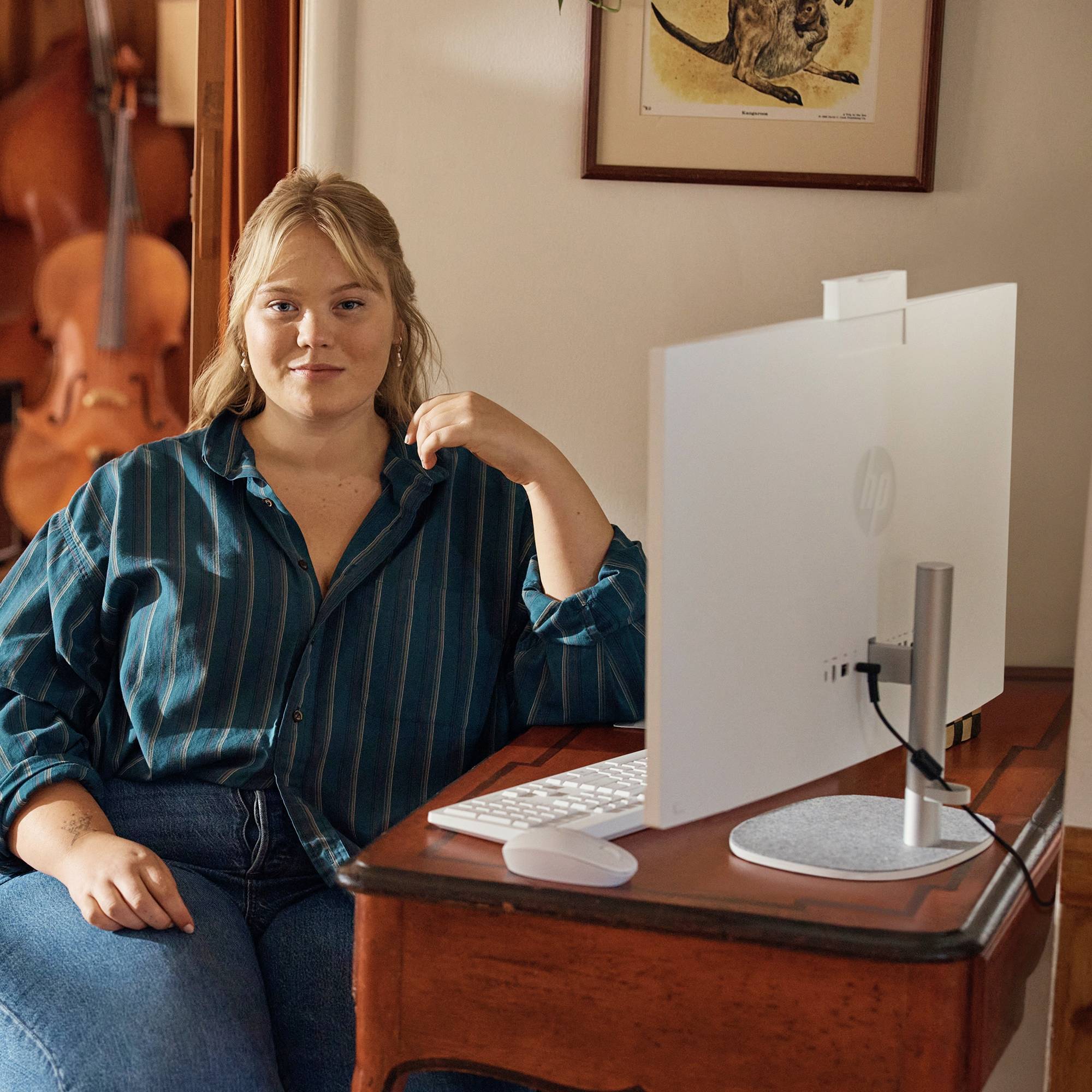 Een vrouw zit aan een bureau naast een HP-desktopmonitor en toetsenbord, poserend in een kamer met een cello op de achtergrond.