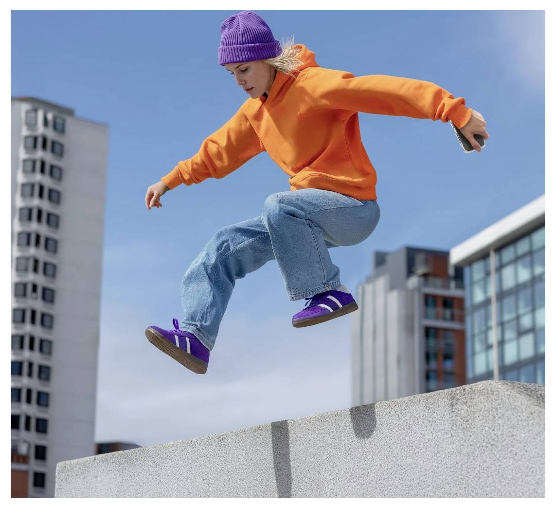 Persoon in oranje hoodie en paarse muts mid-jump over een betonnen rand in een stadssetting.