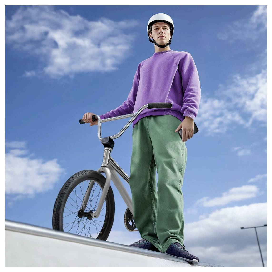 Jonge man in een helm staat met een BMX-fiets op een rand van een skatepark tegen een blauwe hemel.
