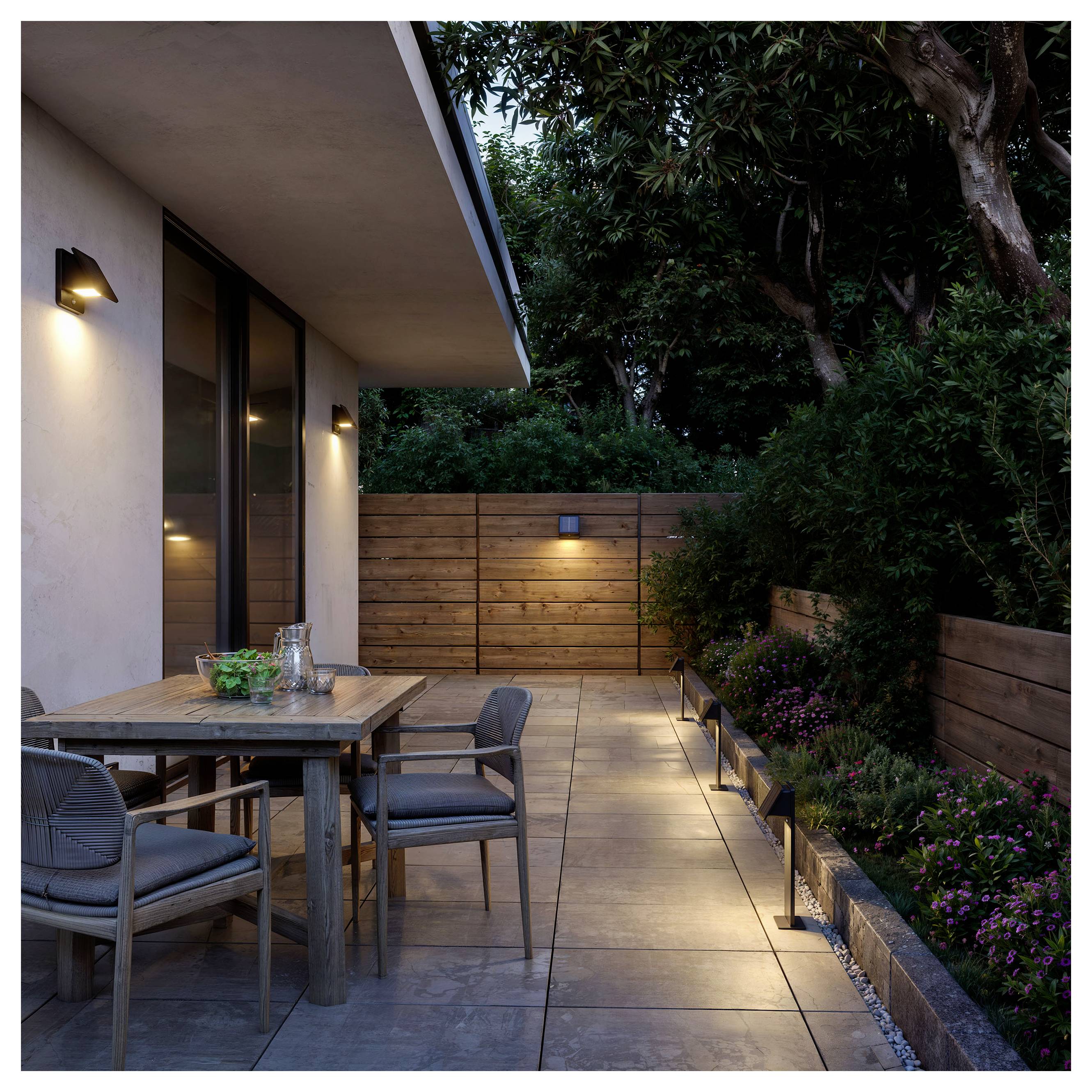 Modern terras bij schemering met een houten eettafel en stoelen naast een huis, verlichte looppad, en aangelegde tuin met bomen en struiken.