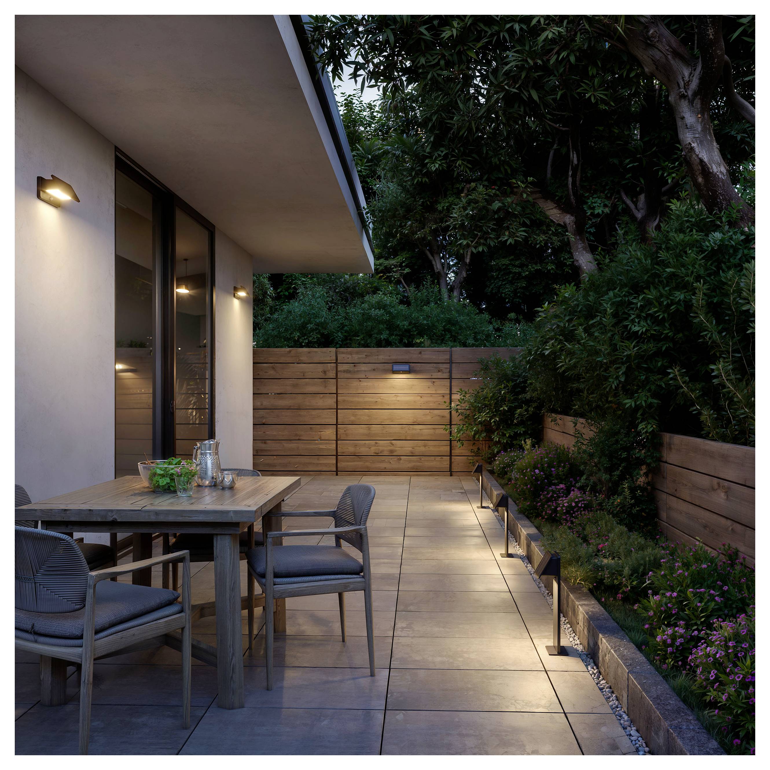 Modern patio bij schemering met buiteneettafel en stoelen naast een huis, verlichte looppad, houten schutting en aangelegde bomen en planten.