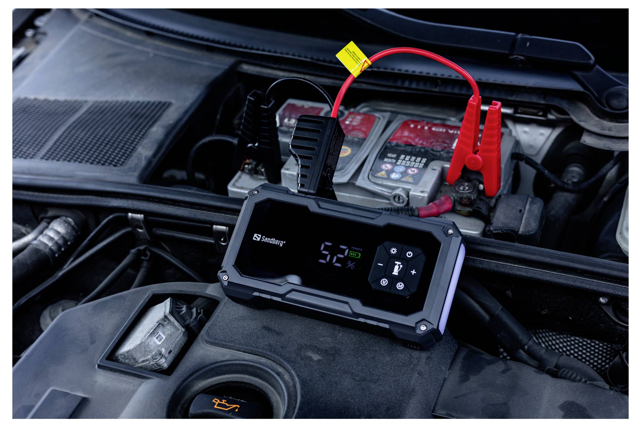 Draagbare jumpstarter aangesloten op een autoaccu in de motorruimte, met rode en zwarte klemmen eraan bevestigd.
