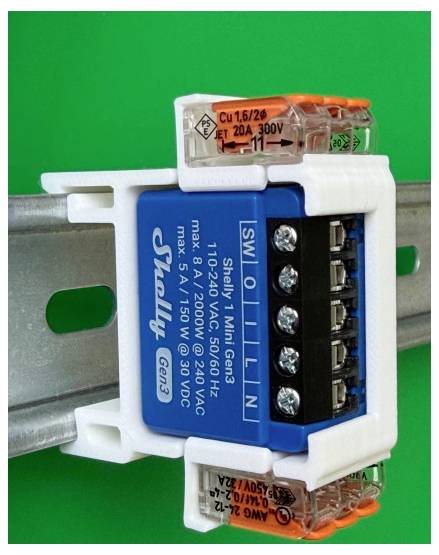 Nahaufnahme van een Shelly 1 Mini Gen3 smart relay-module gemonteerd op een metalen rail, met oranje en transparante draadconnectoren zichtbaar tegen een groene achtergrond.