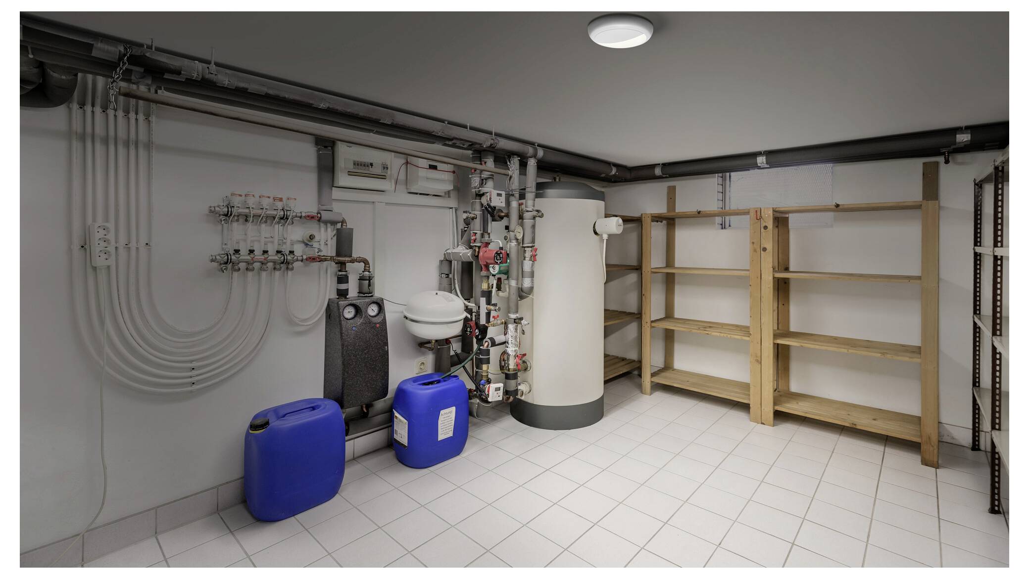 Nutsruimte met waterzuiveringsapparatuur, buizen, een grote tank, blauwe containers en lege houten rekken op een tegelvloer.