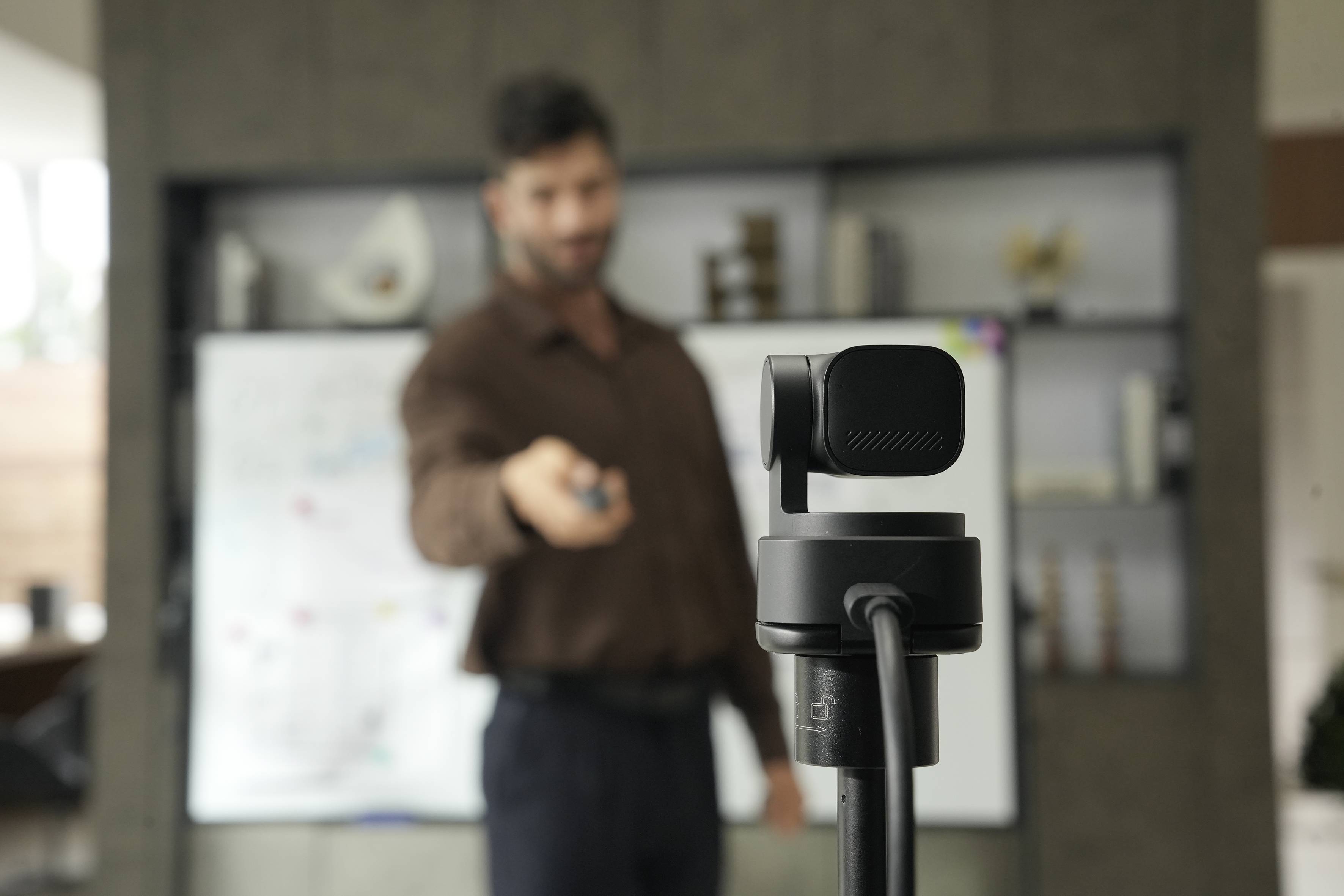 Een webcam op een standaard staat scherp in beeld terwijl een onscherpe man op de achtergrond presenteert, wat suggereert dat er een video wordt opgenomen of een onlinepresentatie plaatsvindt.