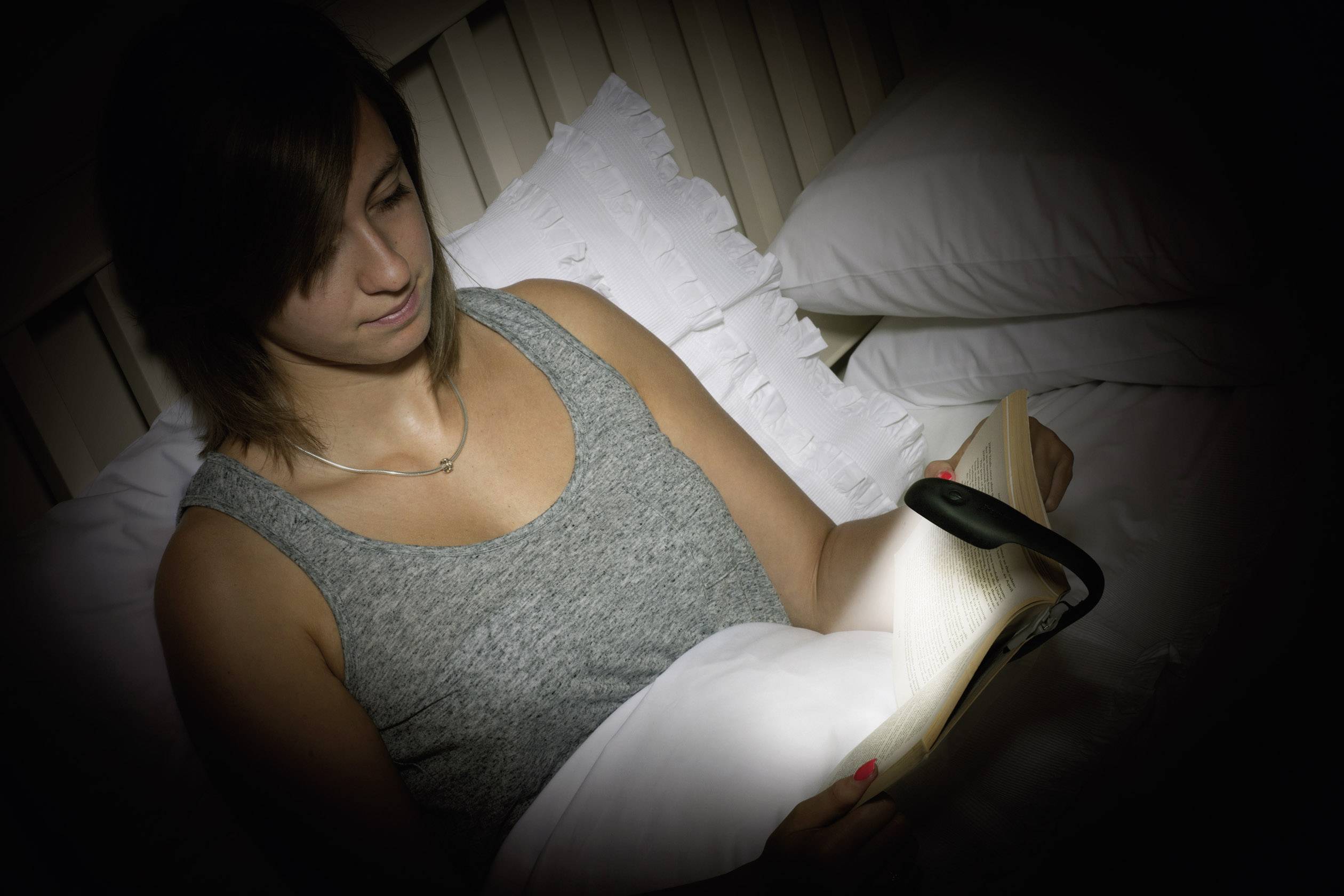 Een persoon in bed leest een boek met een kleine lamp die het boek verlicht. Ze draagt een grijs bovenkleed en ligt half uitgestrekt.