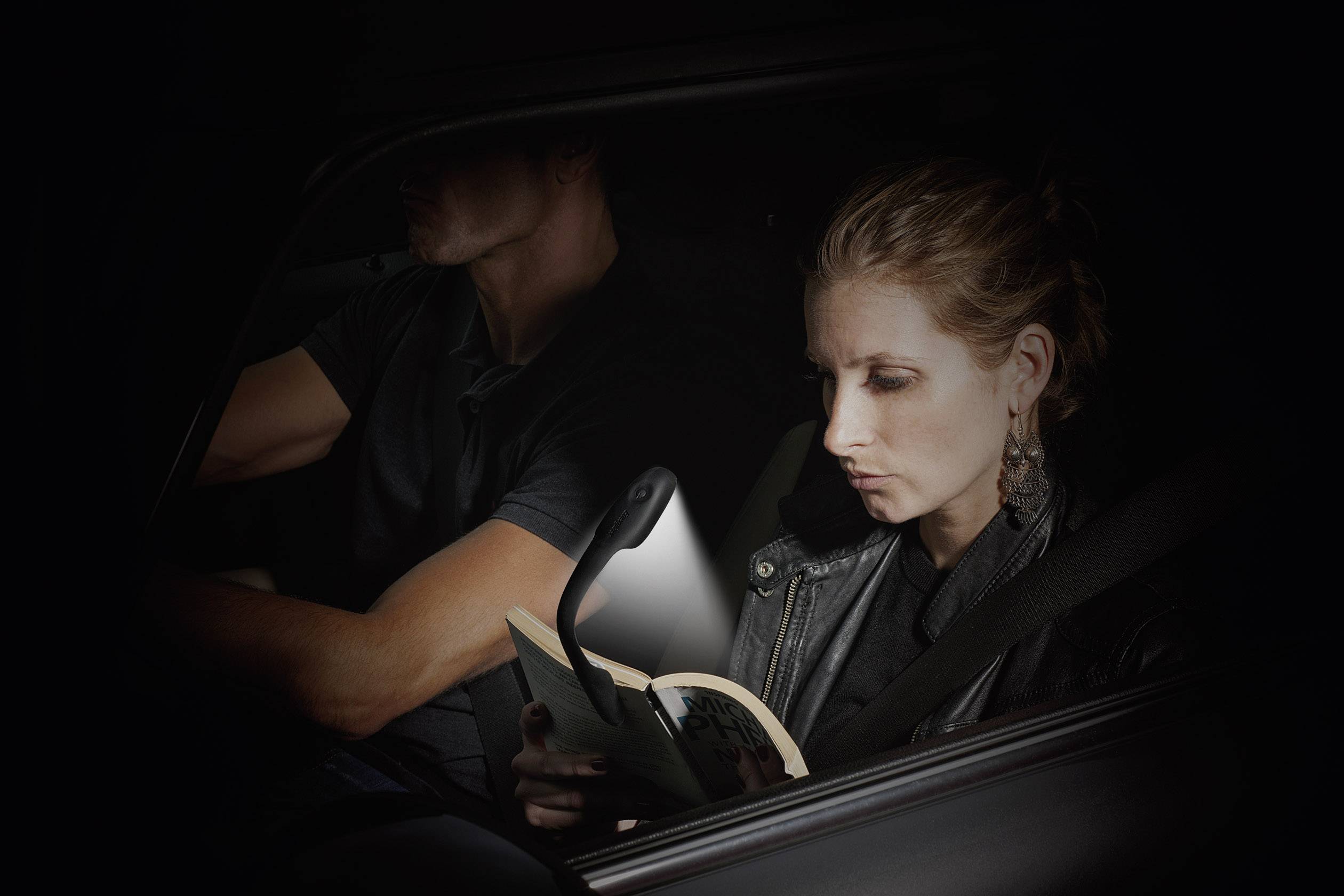 Een vrouw leest een boek in de auto, verlicht door een flexibele leeslamp. Een man zit achter het stuur en rijdt.