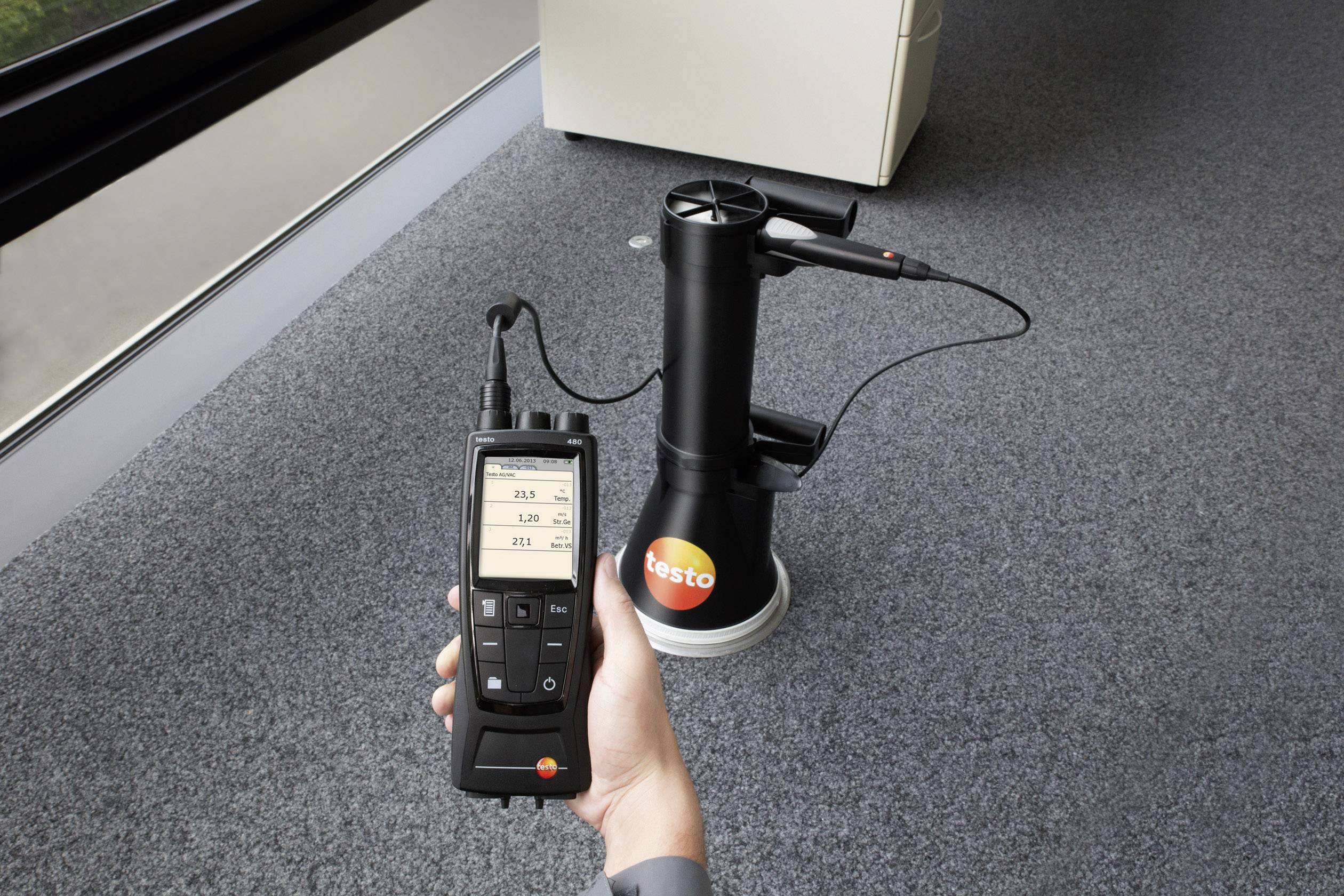 Een handapparaat toont luchtstroommeetwaarden, terwijl een sensor op een tapijtvloer in een kamer staat.
