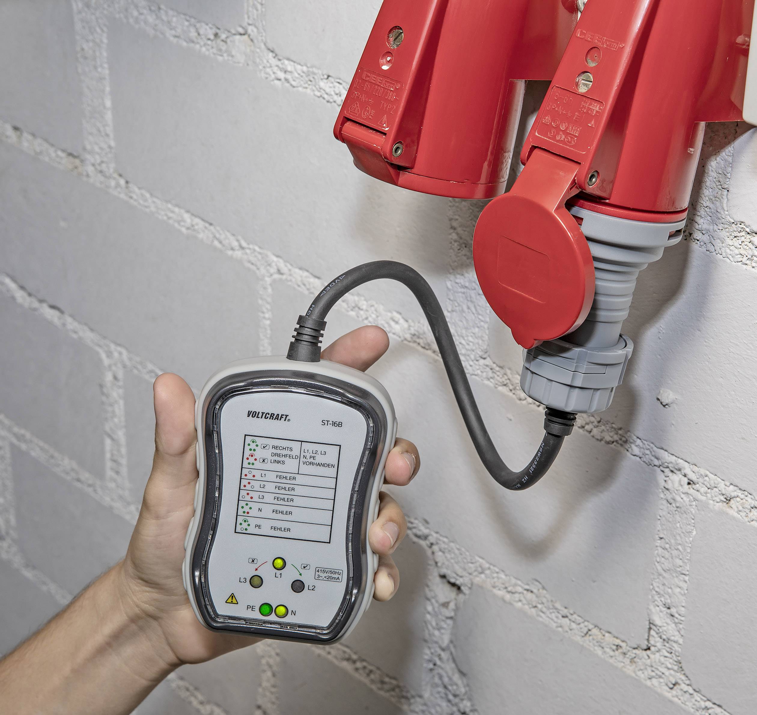 Een hand houdt een meetapparaat vast dat is aangesloten op rode industriële stopcontacten om de elektrische spanning en faseconfiguratie te controleren.