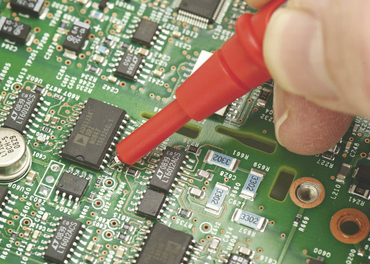 Een hand bedient een rode meetpen op een groene printplaat om elektronische componenten te testen of te meten.