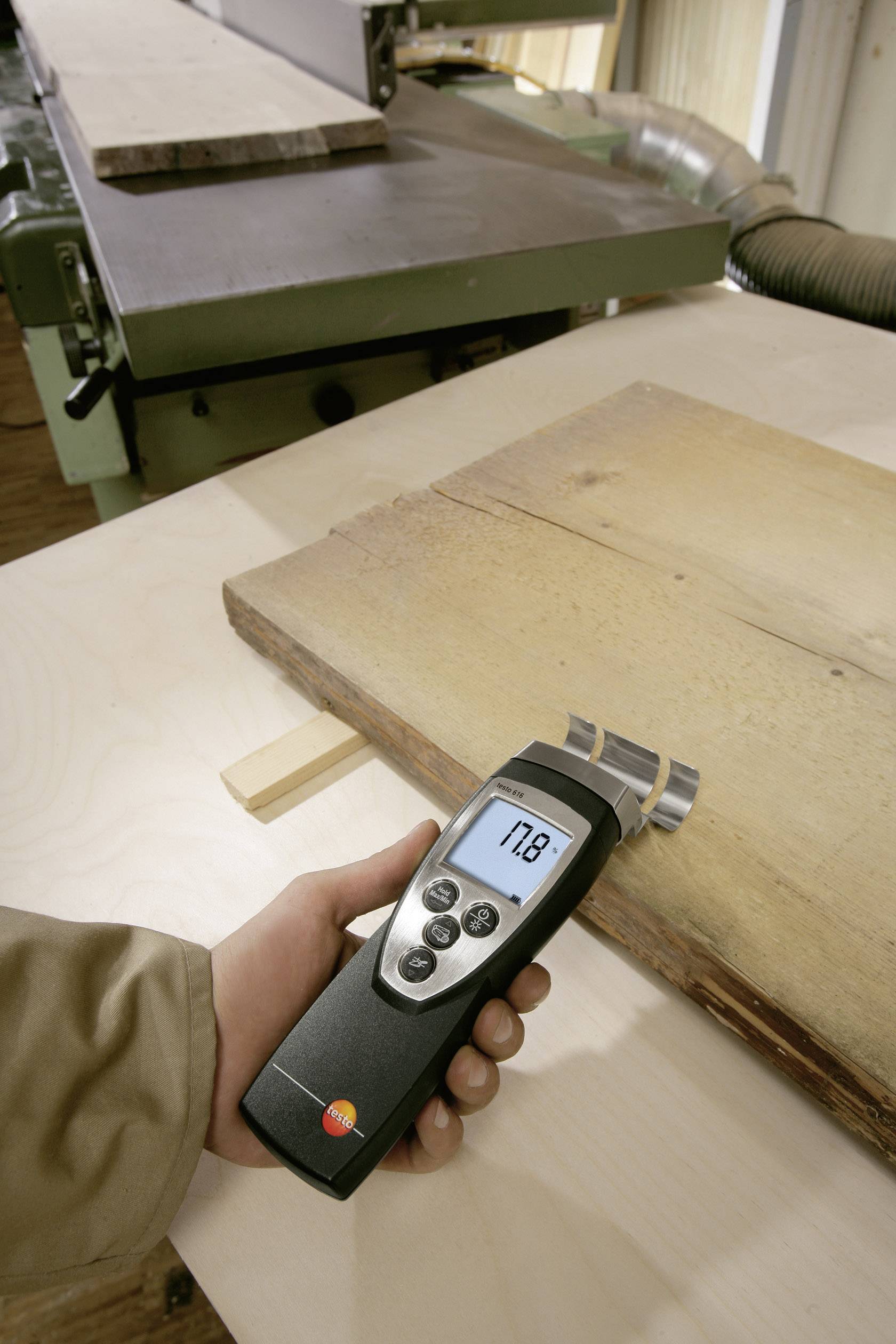 Een persoon meet de vochtigheid van een houten plank met een digitaal meetapparaat in een werkplaats, met een machine op de achtergrond rechts.