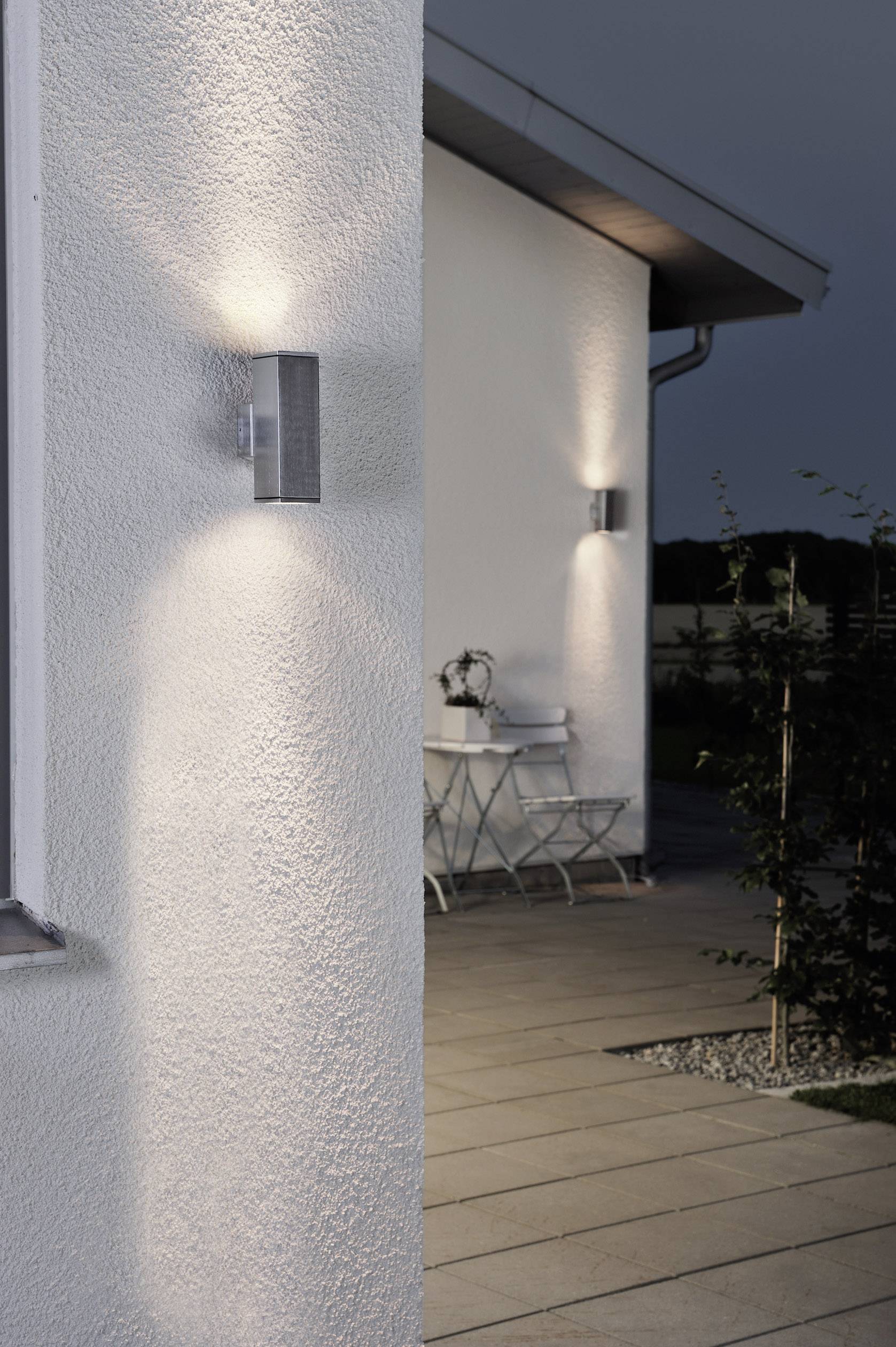 Moderne buitenverlichting aan een witte muur verlicht de ingang. Op de achtergrond bevindt zich een terras met tuinmeubels.