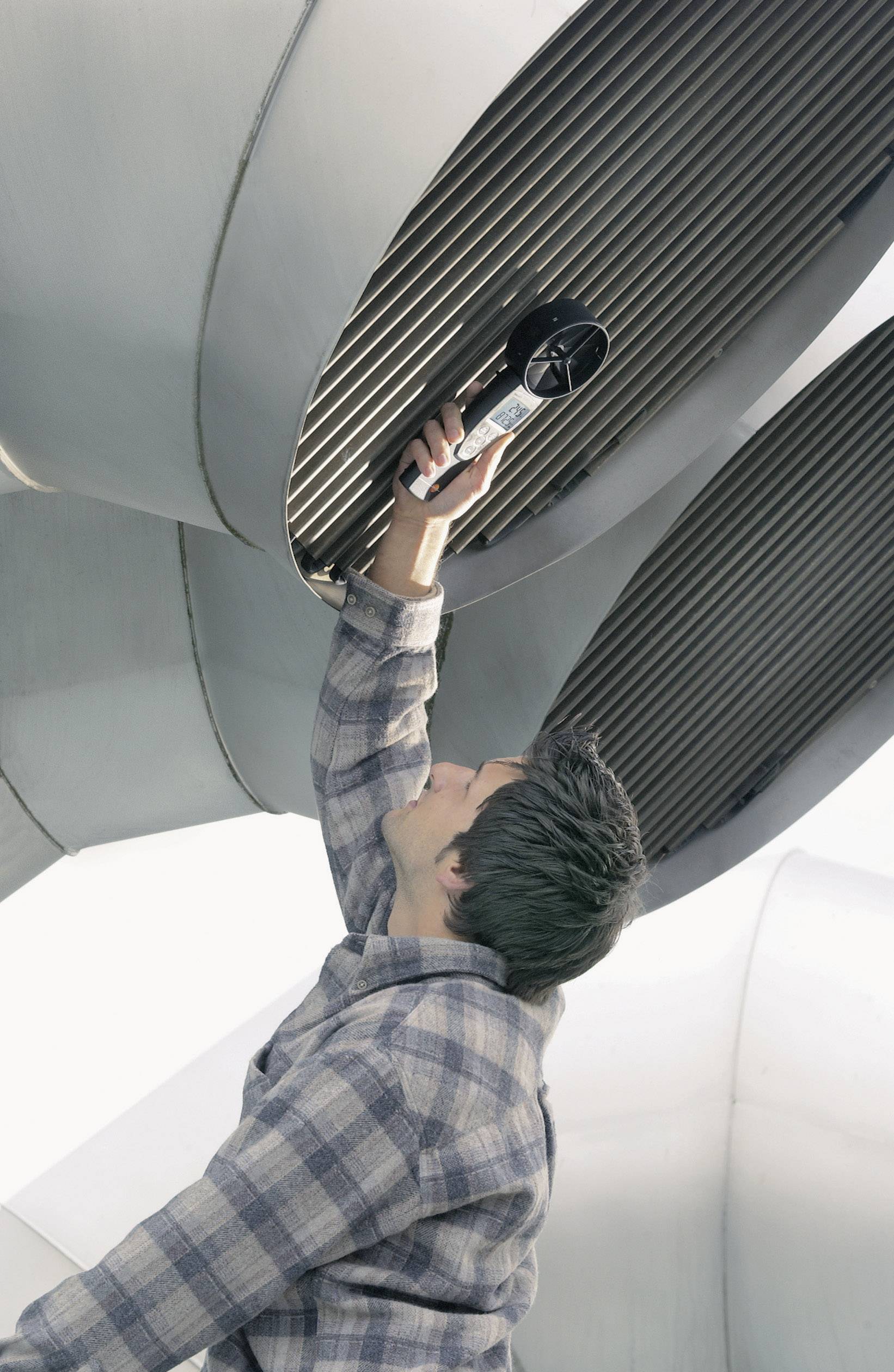 Man in geruit overhemd controleert grote ventilatieopeningen met een apparaat in een industriële omgeving, waarschijnlijk tijdens onderhoudswerkzaamheden.