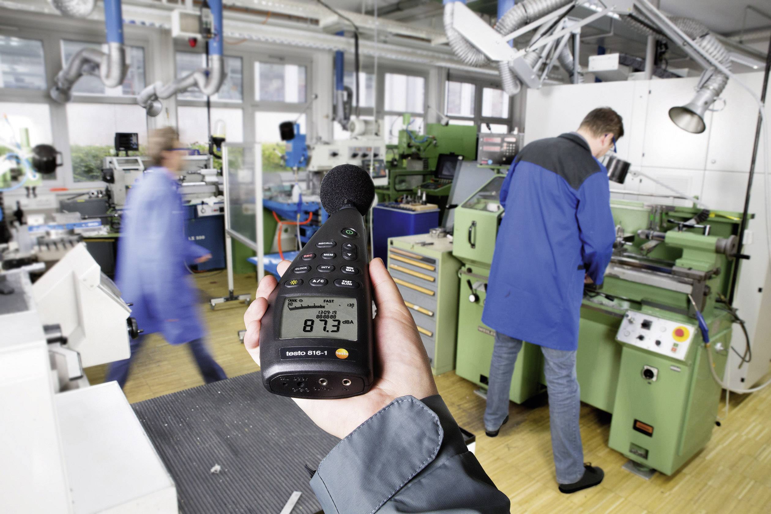 Een persoon houdt een geluidsniveaumeter vast in een werkplaats, terwijl twee personen aan machines werken. De meetwaarde toont 87,3 dB.