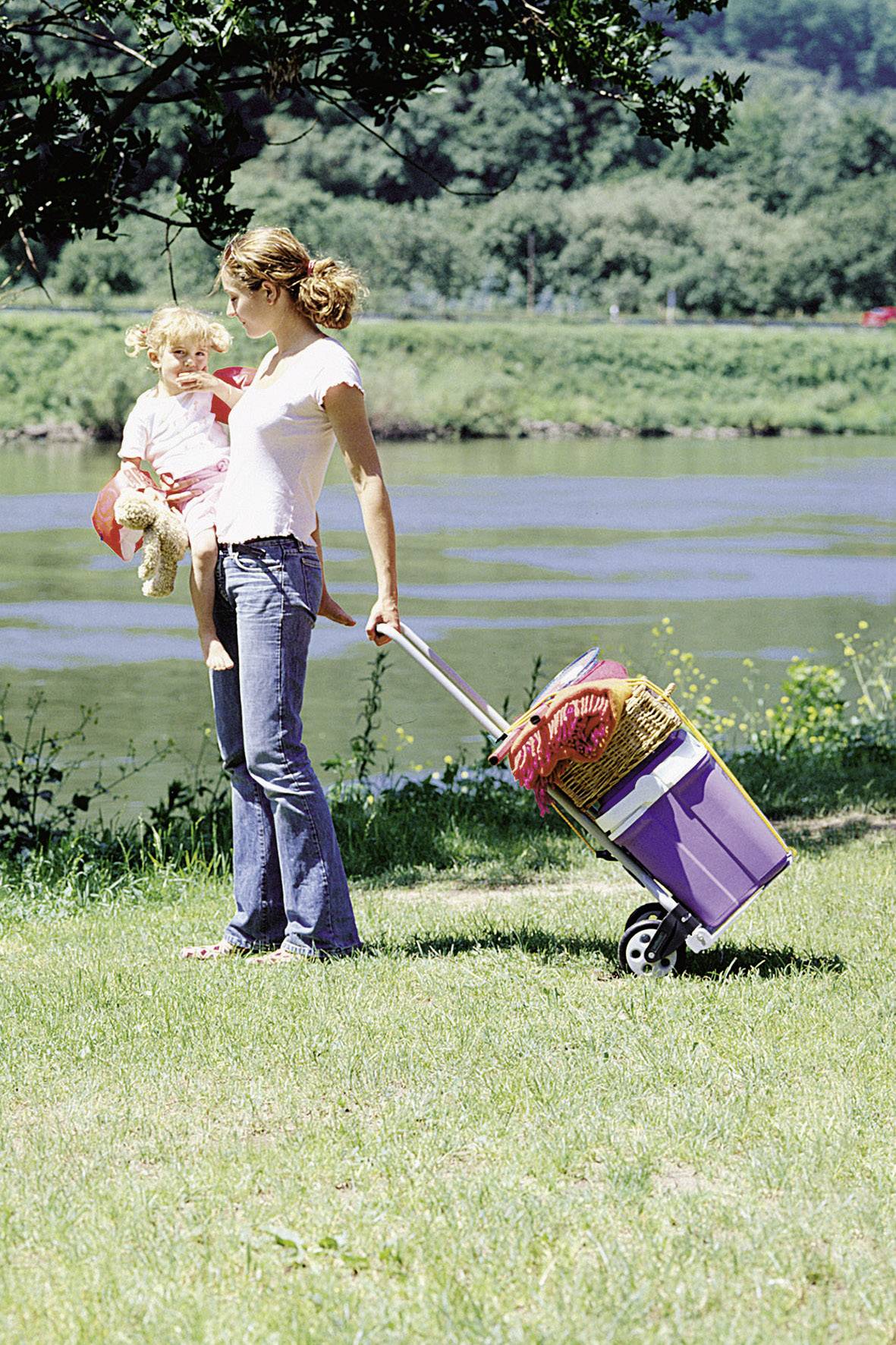 Een vrouw in jeans en een wit T-shirt trekt een paarse wagen met picknickmand. Een klein kind met een knuffeldier zit op haar arm.