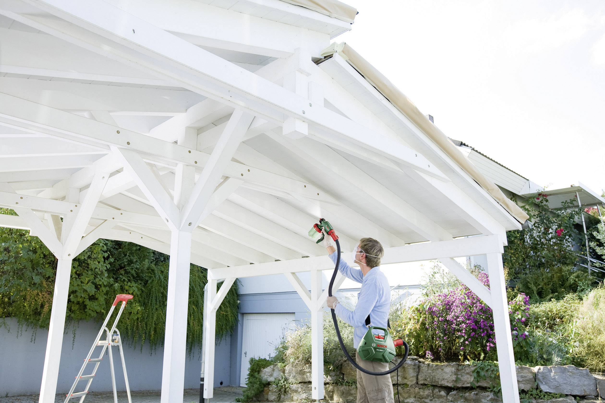 Een persoon spuit het witte houten frame van een carport buiten met een verfspuitapparaat. Een ladder staat ernaast.