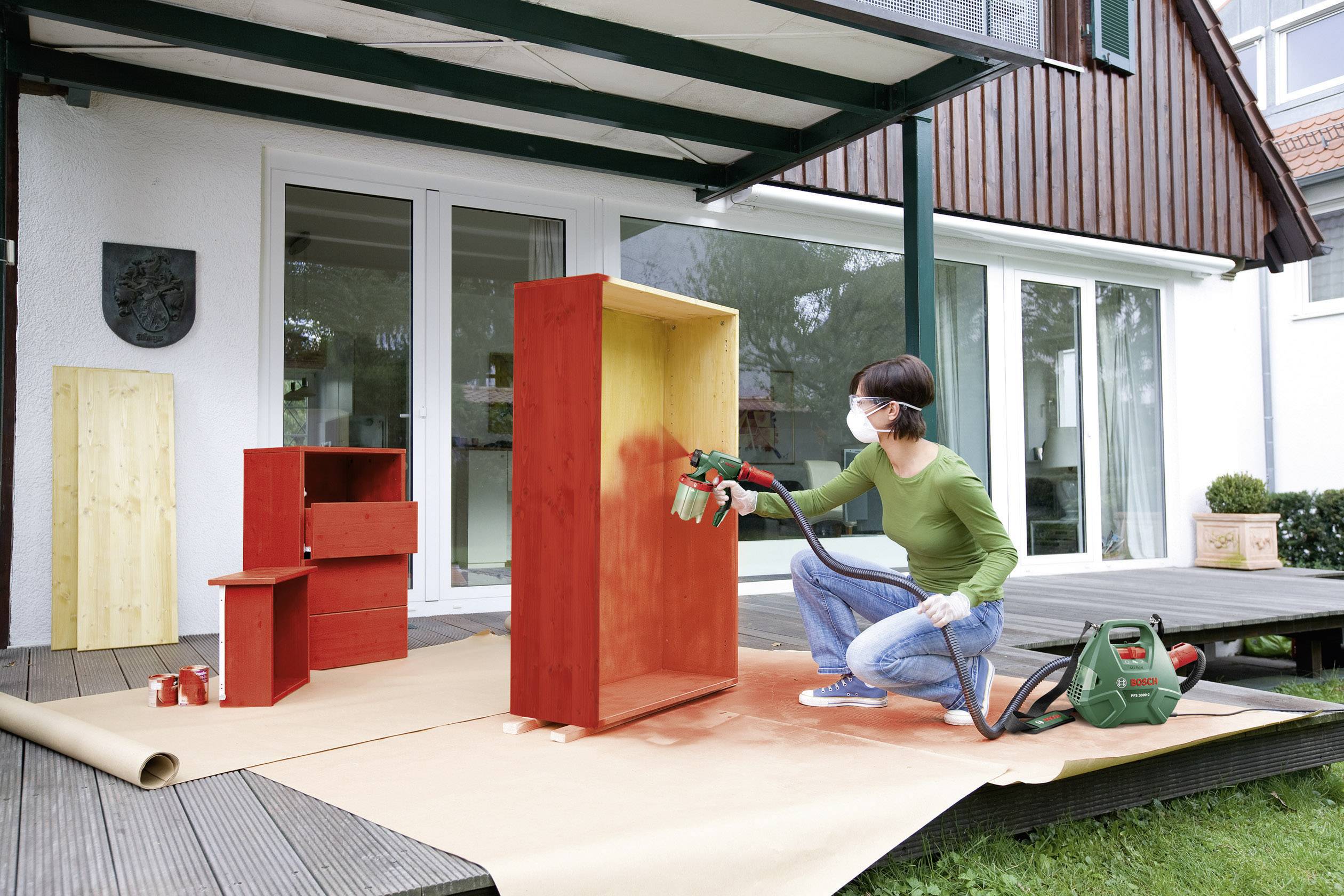 Een persoon spuit rode verf op een houten rek met een verfspuitpistool op een terras. Andere geschilderde meubelstukken zijn op de achtergrond zichtbaar.