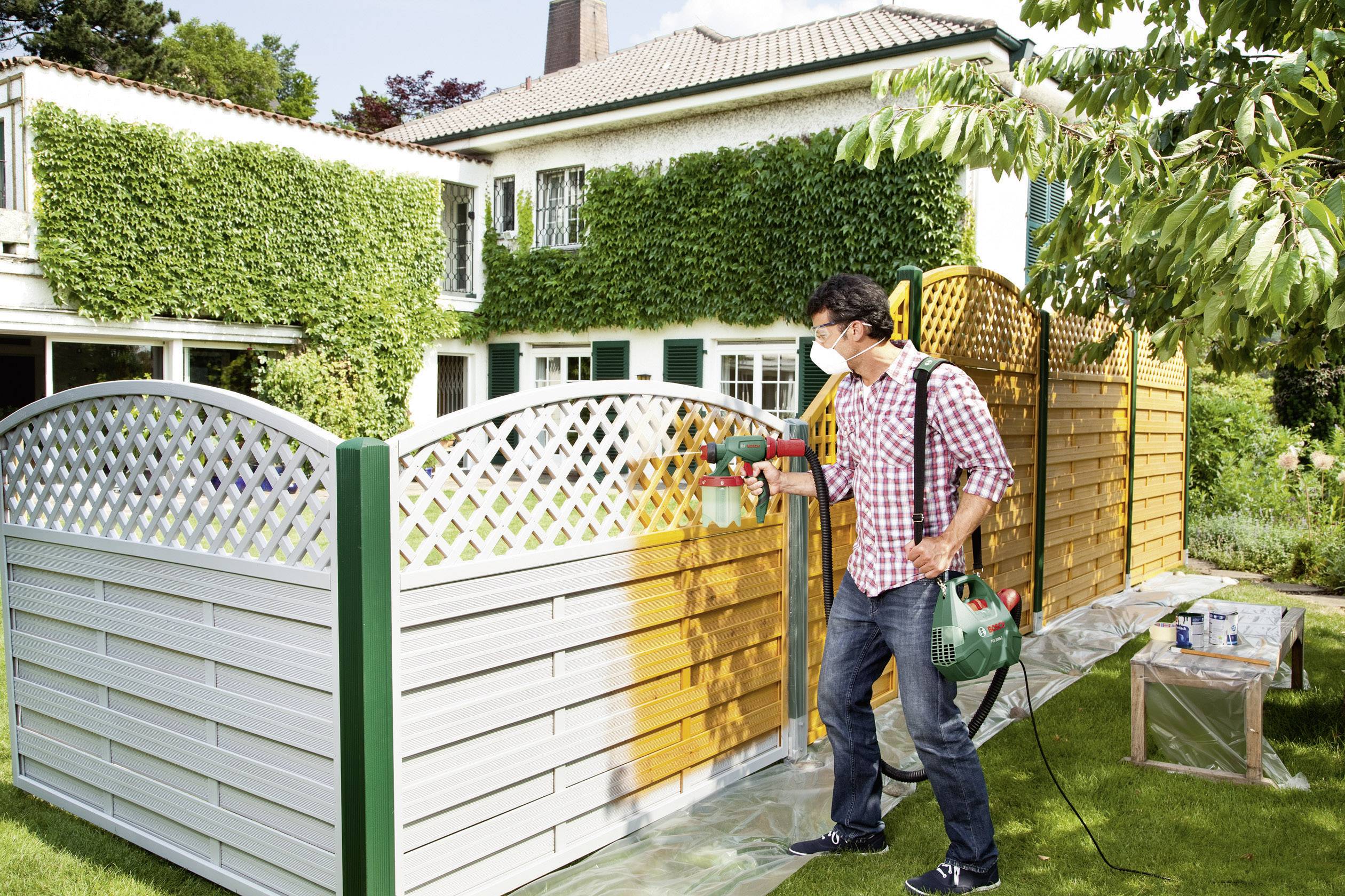 Een man draagt een beschermingsmasker en schildert een houten hek met een verfspuitpistool voor een met klimop begroeid huis.