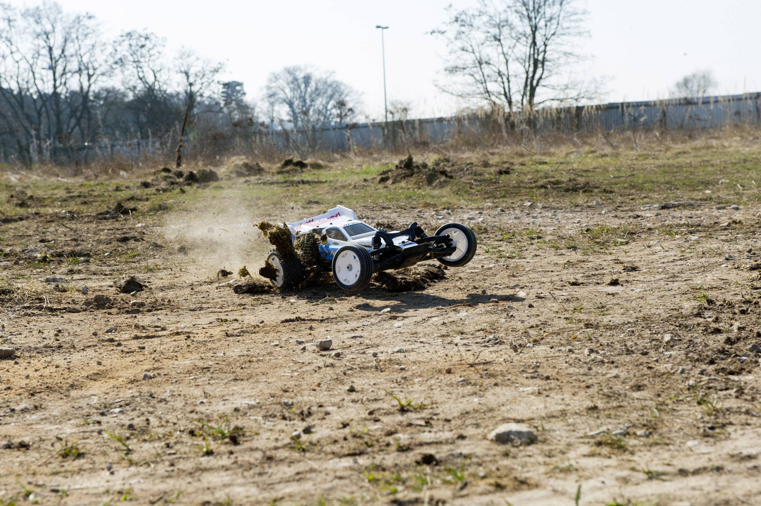 Een bestuurbare auto rijdt snel over een stoffig, kaal landschap en werkt aarde op. Op de achtergrond staan kale bomen.