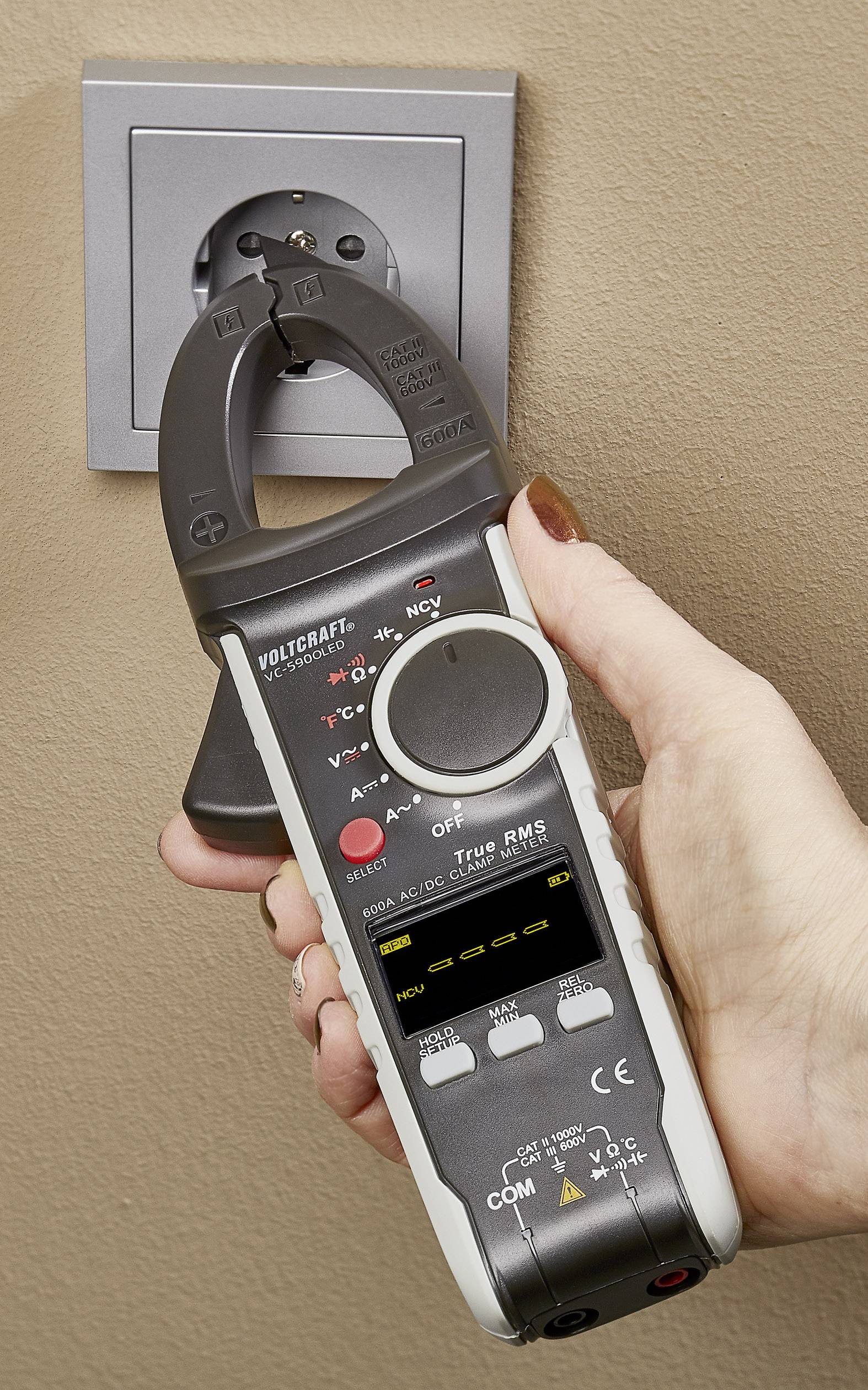 Een hand houdt een digitale stroommeterset vast, die klemmen sluit rond een stroomkabel boven een stopcontact. Het apparaat toont meetwaarden.