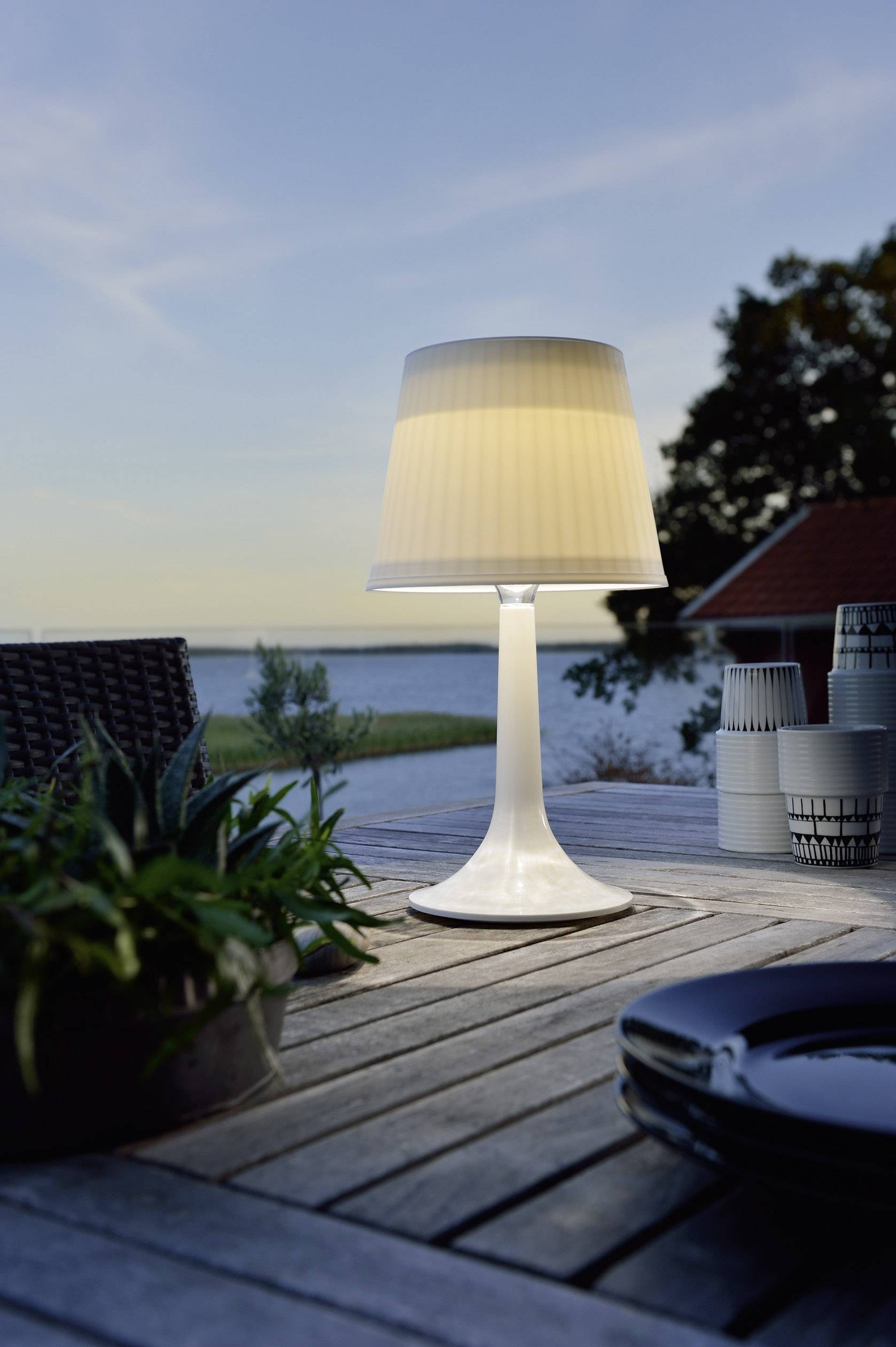 Een tafellamp schijnt op een houten tafel buiten. Op de achtergrond zijn een meer en een bewolkte lucht zichtbaar.