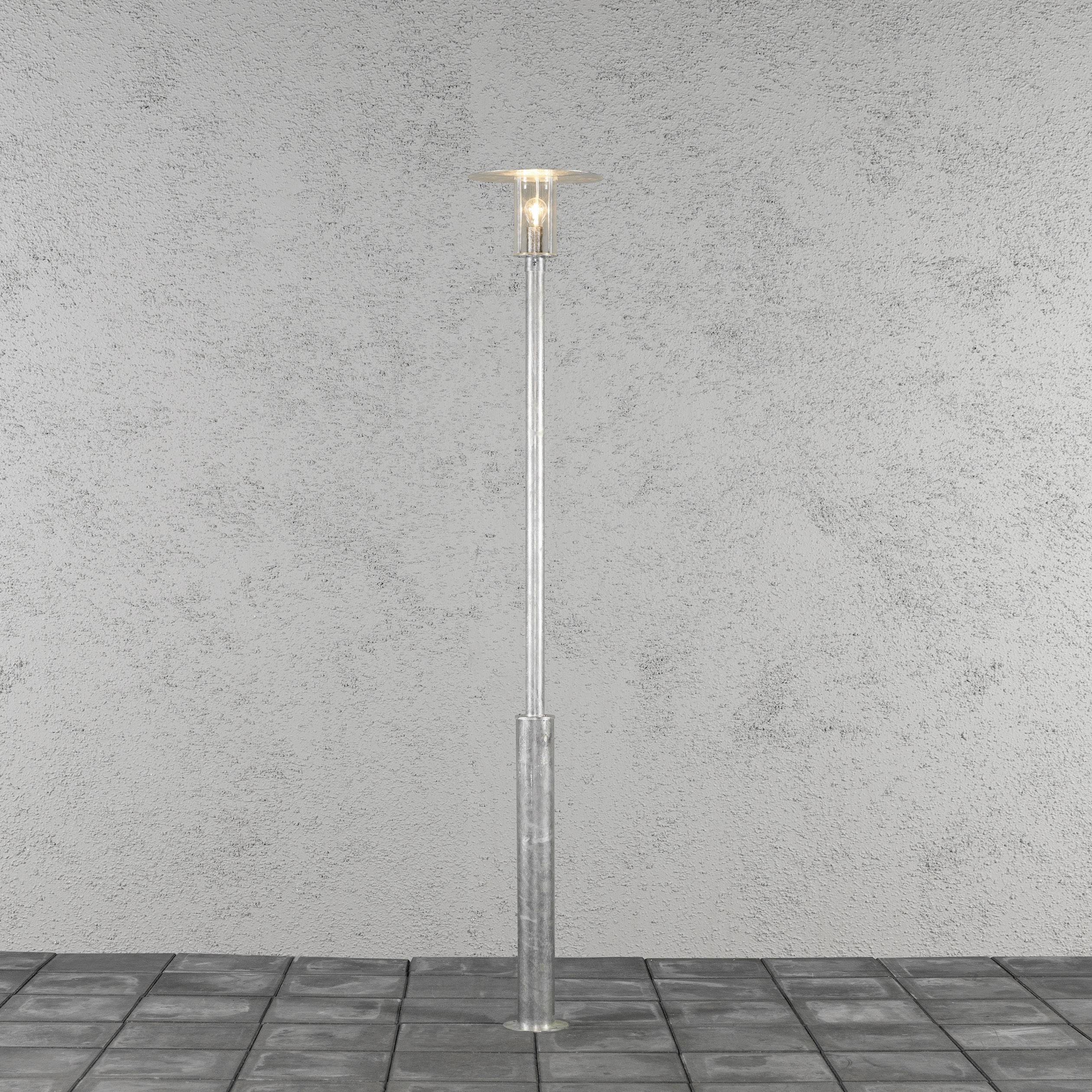 Een eenvoudige, zilverkleurige staande lamp met een cilindrische voet en een glazen lampenkap staat op een grijze betegelde vloer.