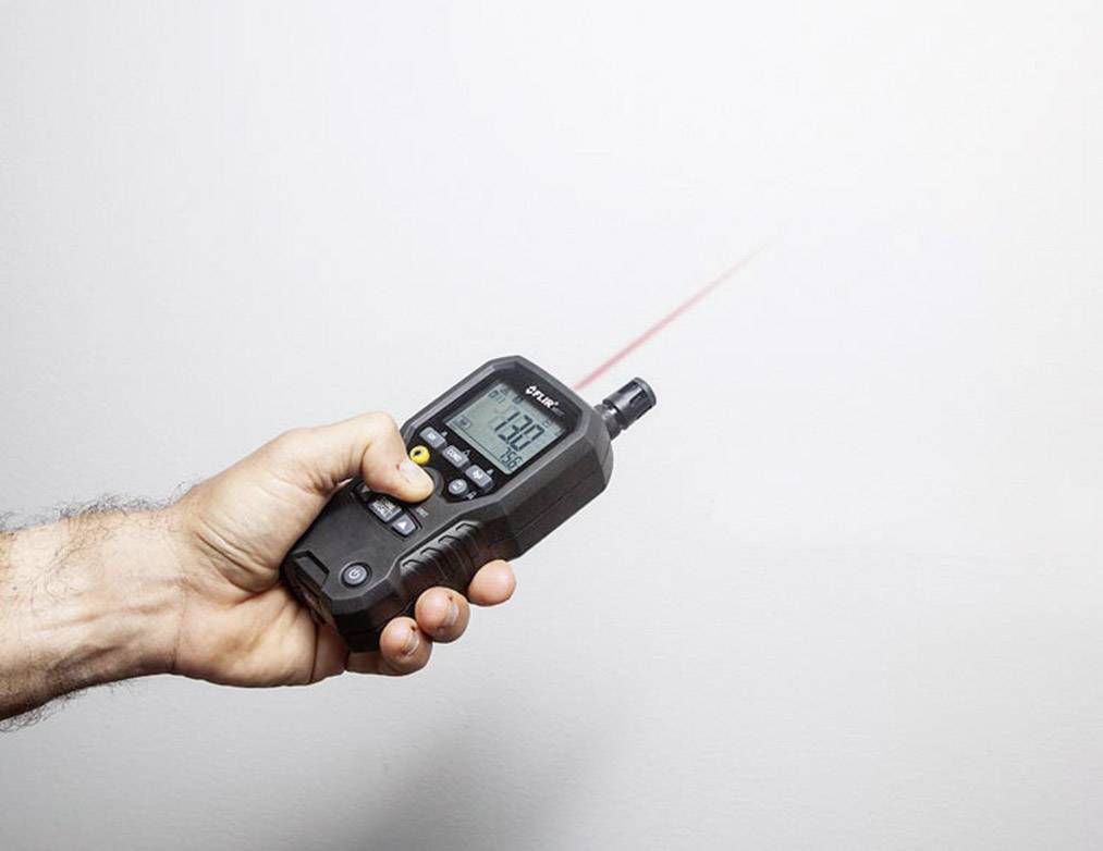 Een hand houdt een laserafstandsmeter vast, gericht op een muur. Het display toont een meting van '129'.