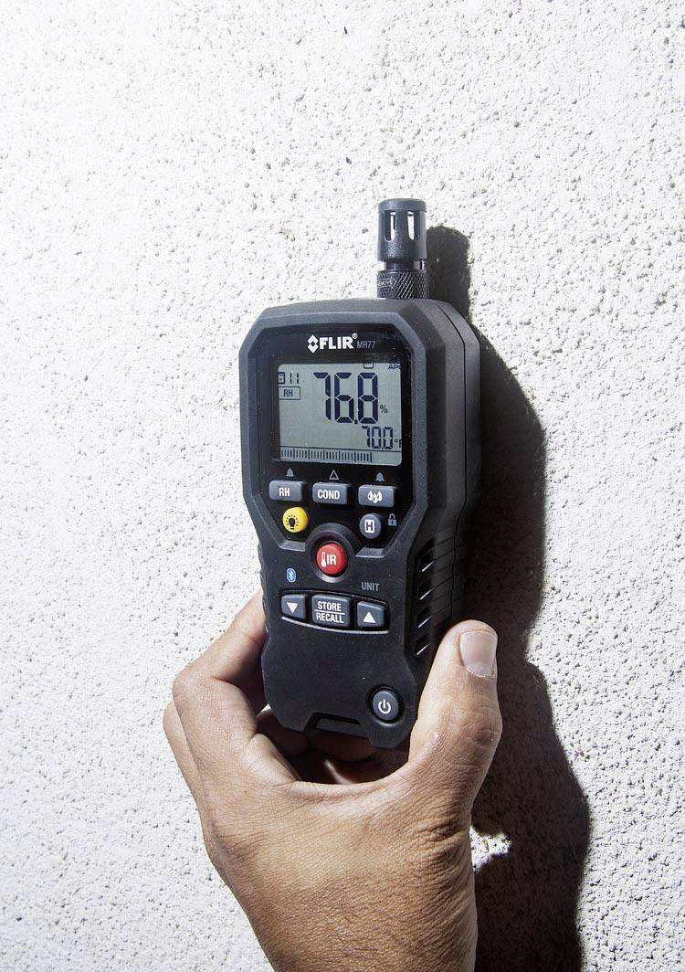 Een hand houdt een digitaal meetapparaat tegen een muur, toont een temperatuur van 76,8 °F. Het apparaat biedt temperatuur- en vochtmetingen.