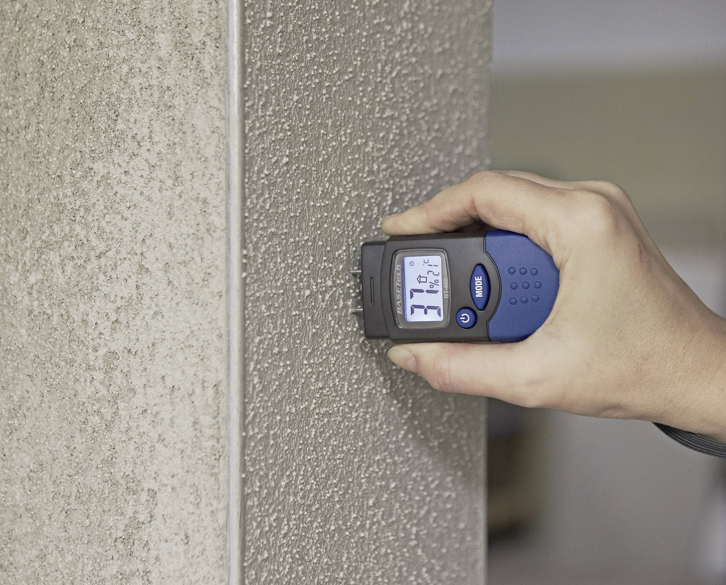 Een hand houdt een elektrische vochtmeter tegen een betonnen muur. Het display toont het vochtgehalte van de muur.
