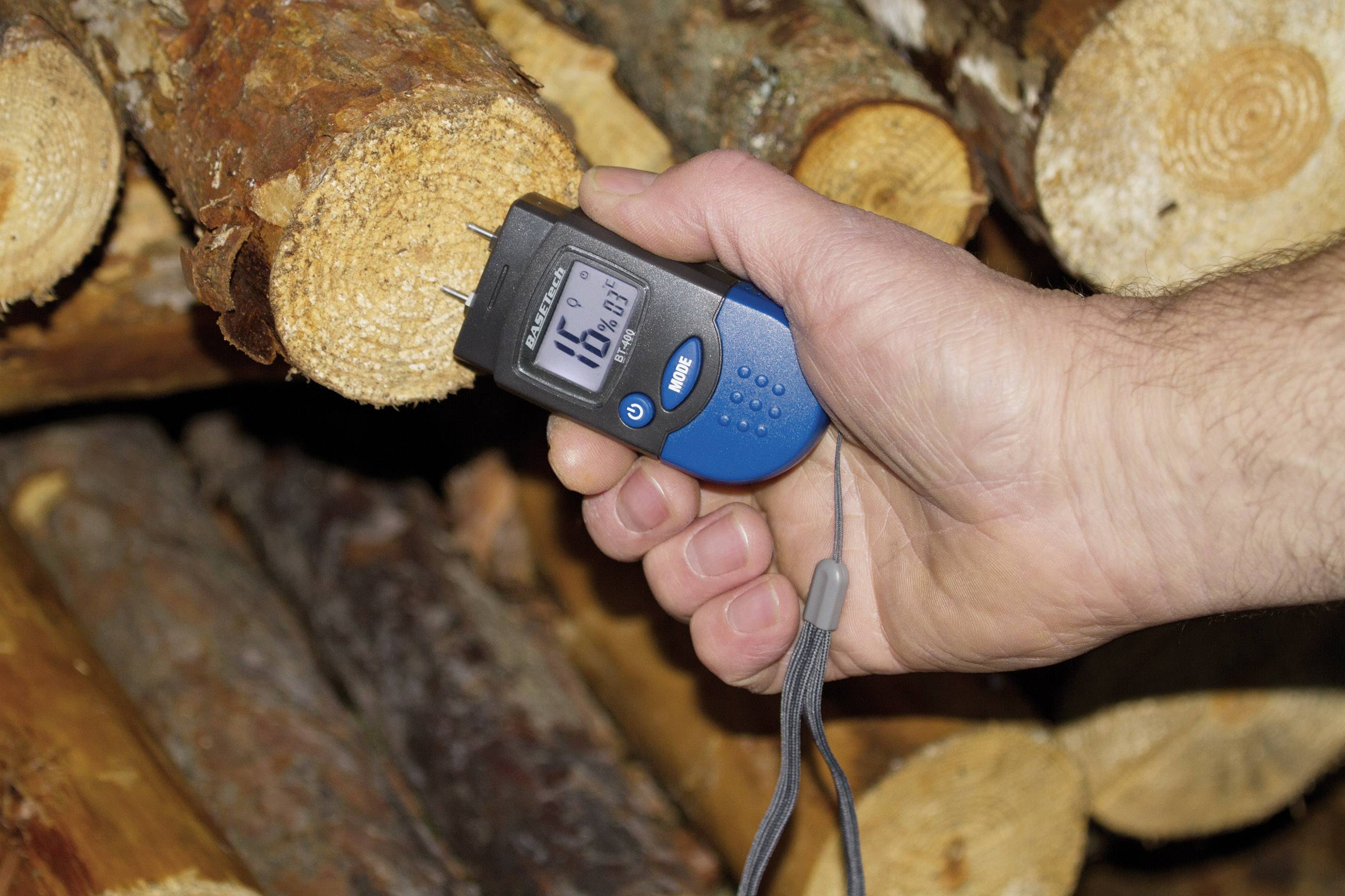 Een hand houdt een vochtmeter tegen een stuk gezaagd hout, waarbij het scherm '16,3%' aangeeft. Op de achtergrond: gestapelde brandhoutblokken.