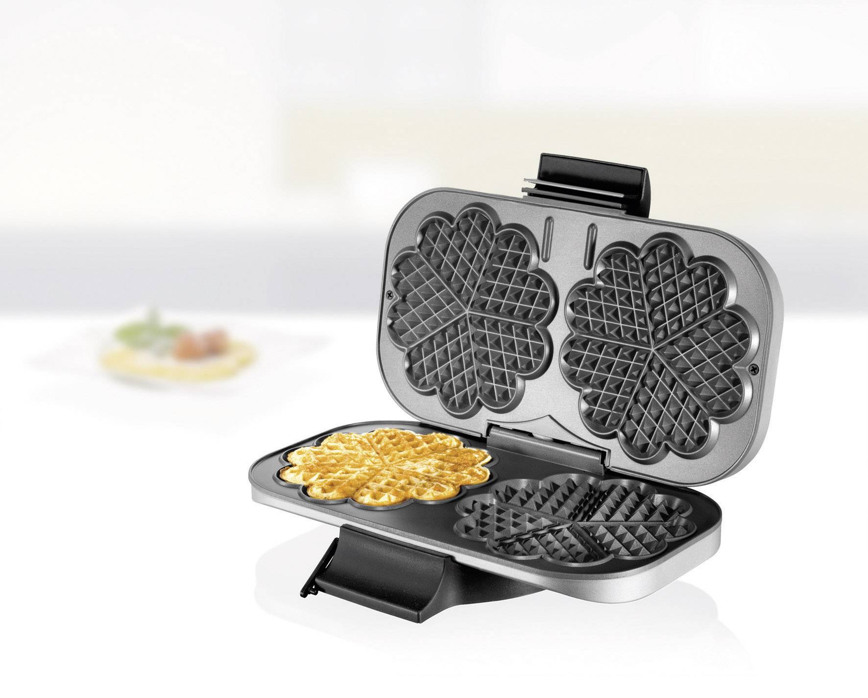 Een wafelijzer met een open deksel dat een hartvormige wafel toont. Op de achtergrond een bord met een wafel en decoratie.