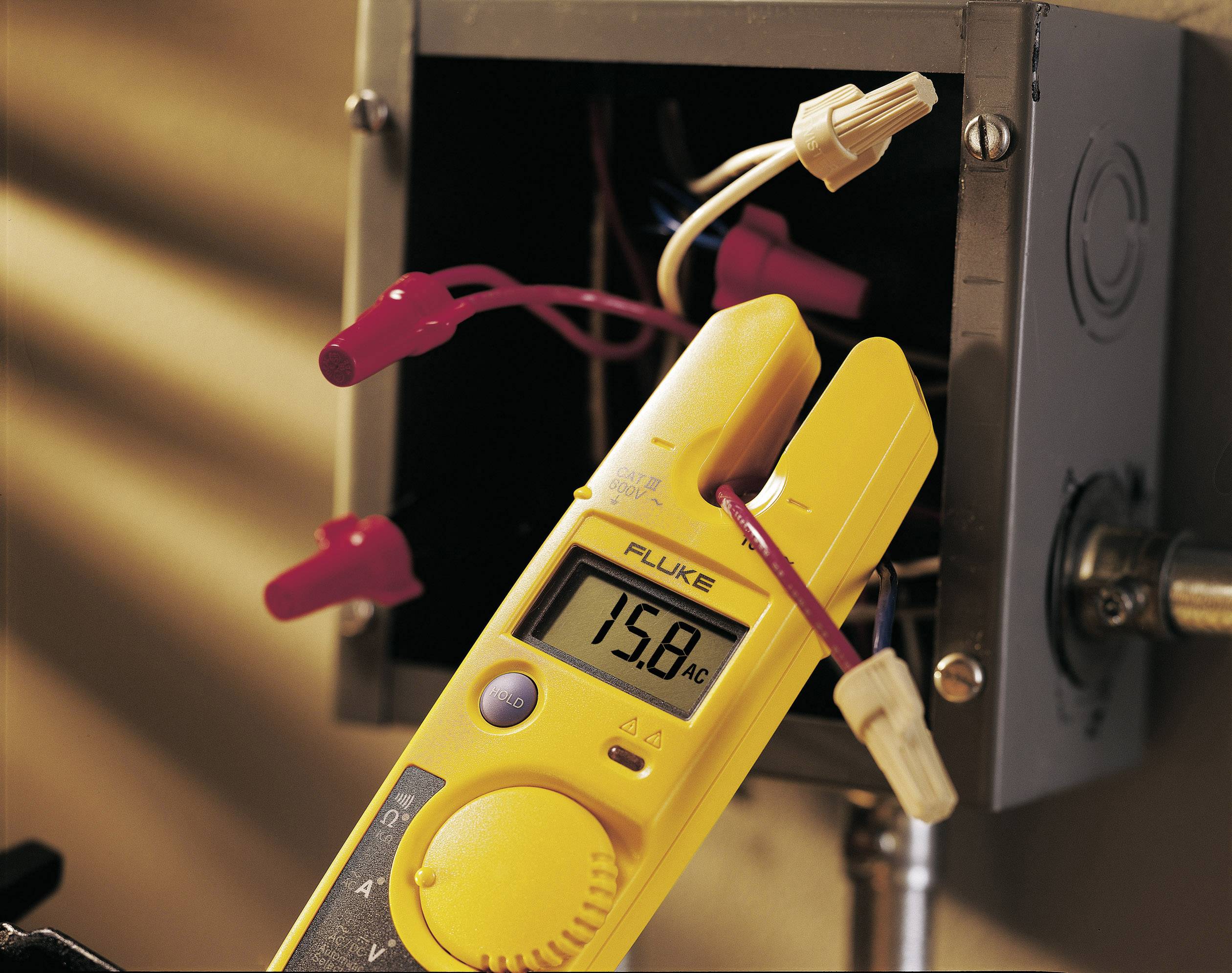 Een gele multimeter meet 15,8 ampère wisselstroom aan een geopende aansluitkast met zichtbare draden en verbindingen.