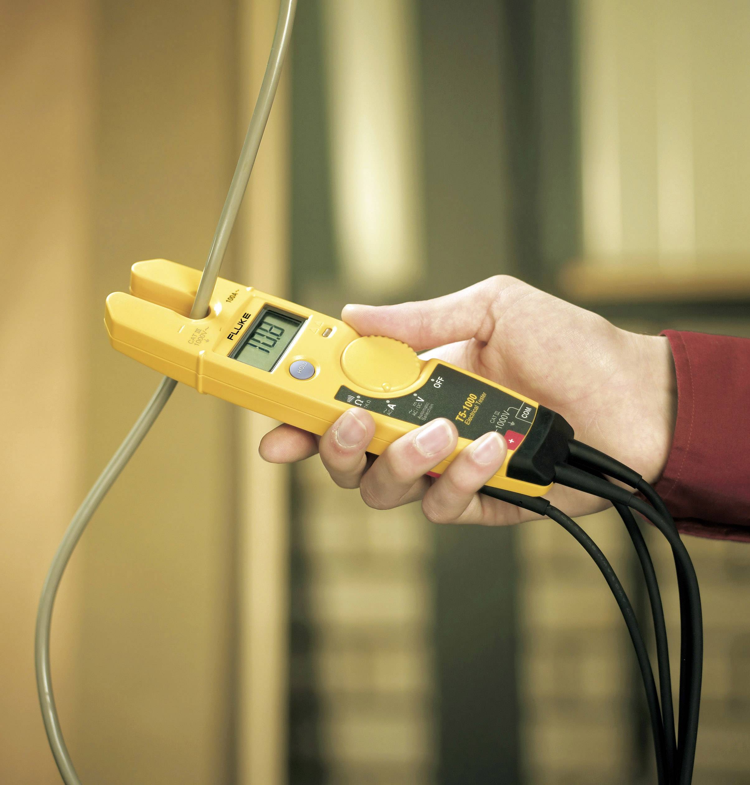 Een hand houdt een geel elektrisch meettoestel vast dat een spanning van 120,2 volt aangeeft en is aangesloten met kabels.