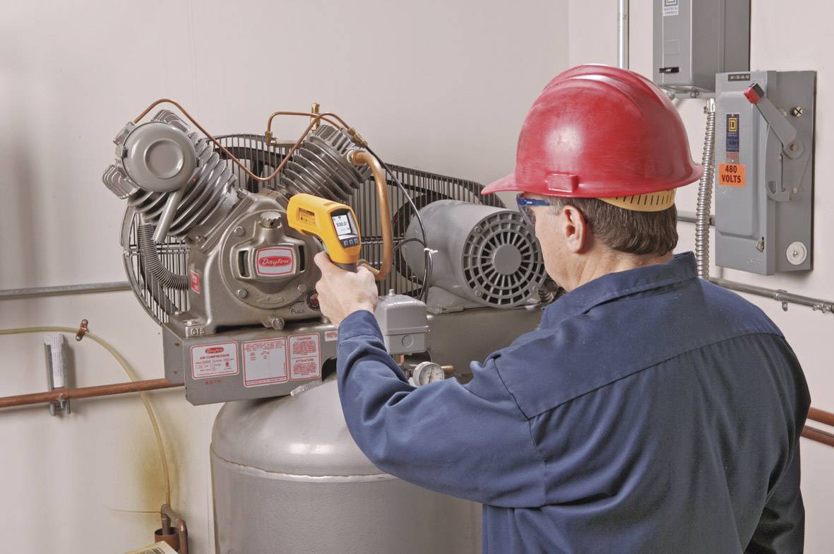 Een man met een veiligheidshelm en werkkleding controleert een industriële compressor met een infrarood-thermometer.