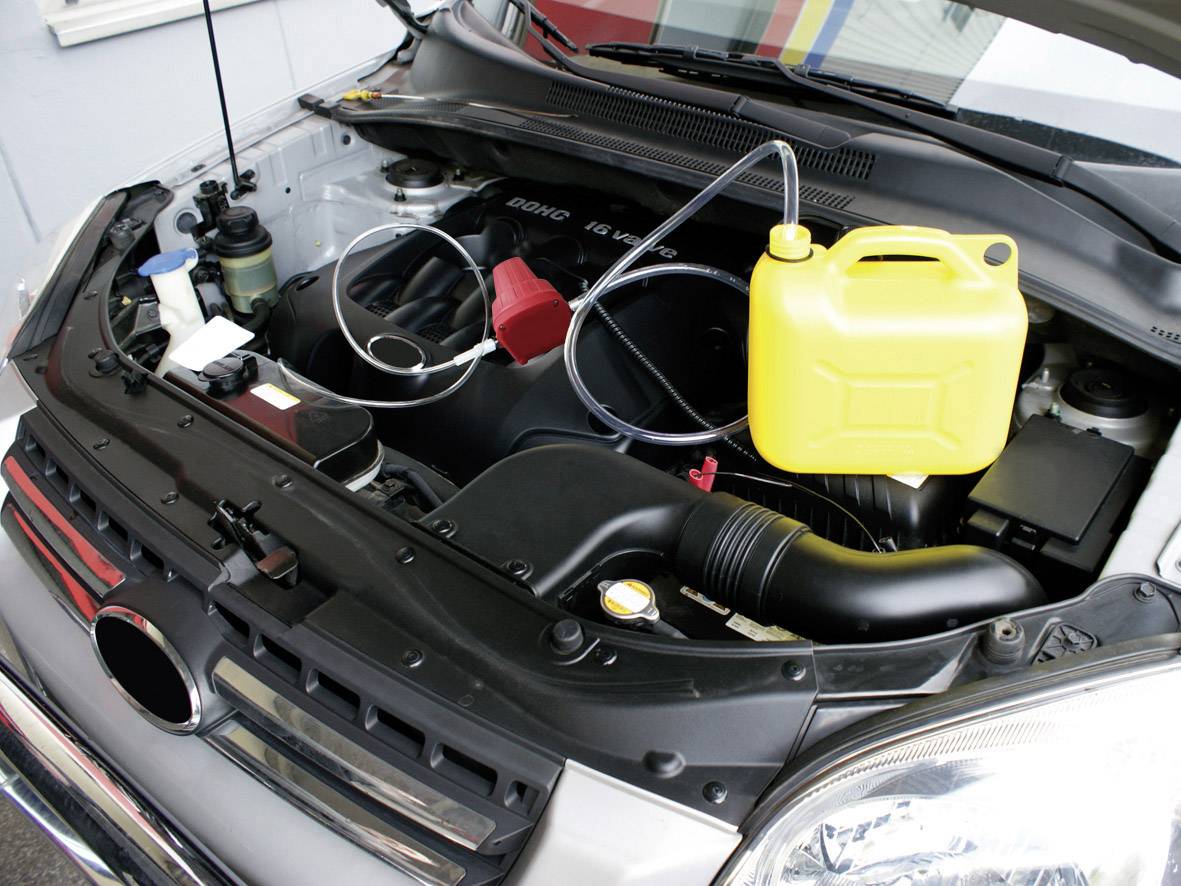 Een geopende motorruimte met een gele jerrycan op het motorblok en slangen die met de jerrycan zijn verbonden, toont een koelvloeistofvulsysteem.